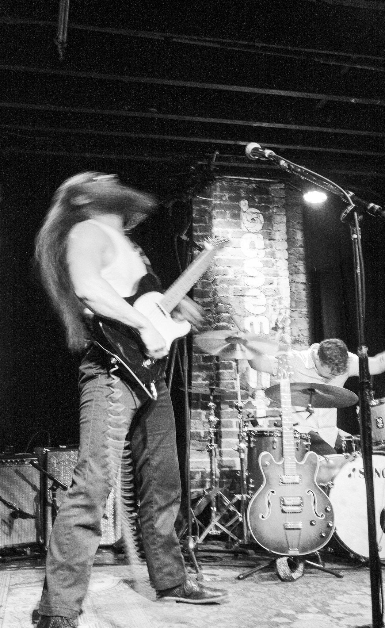 A black and white photo of a musician playing an electric guitar on stage with a drummer in the background and a brick wall behind them.
