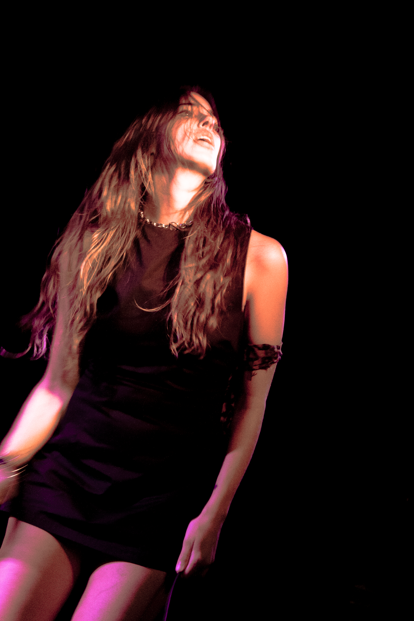 A woman with long hair, wearing a black sleeveless dress, singing passionately on stage with a black background.