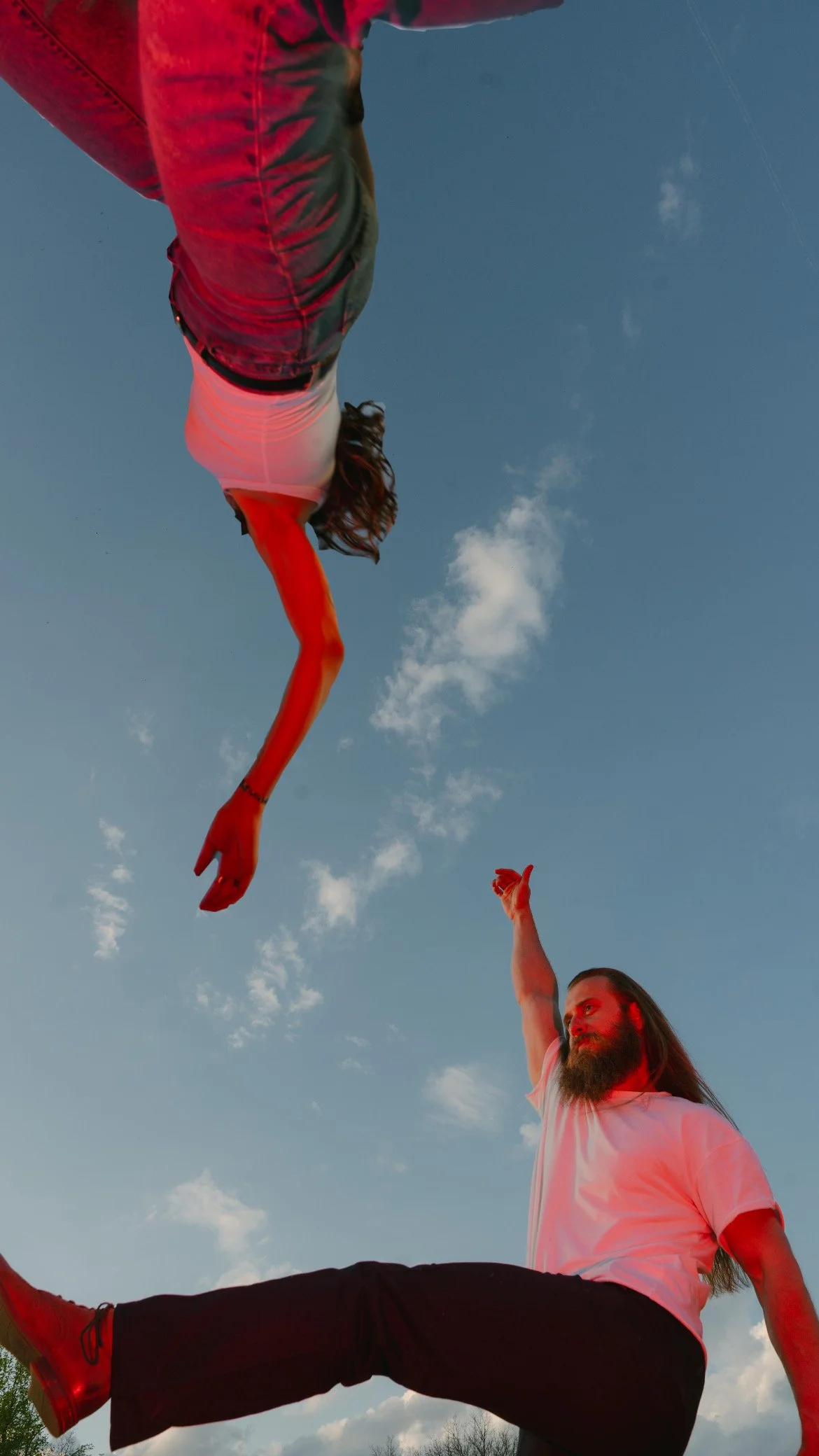 A man with a beard and long hair is sitting on a ledge outdoors during the daytime, raising one arm while a woman is floating above him, extending one arm.