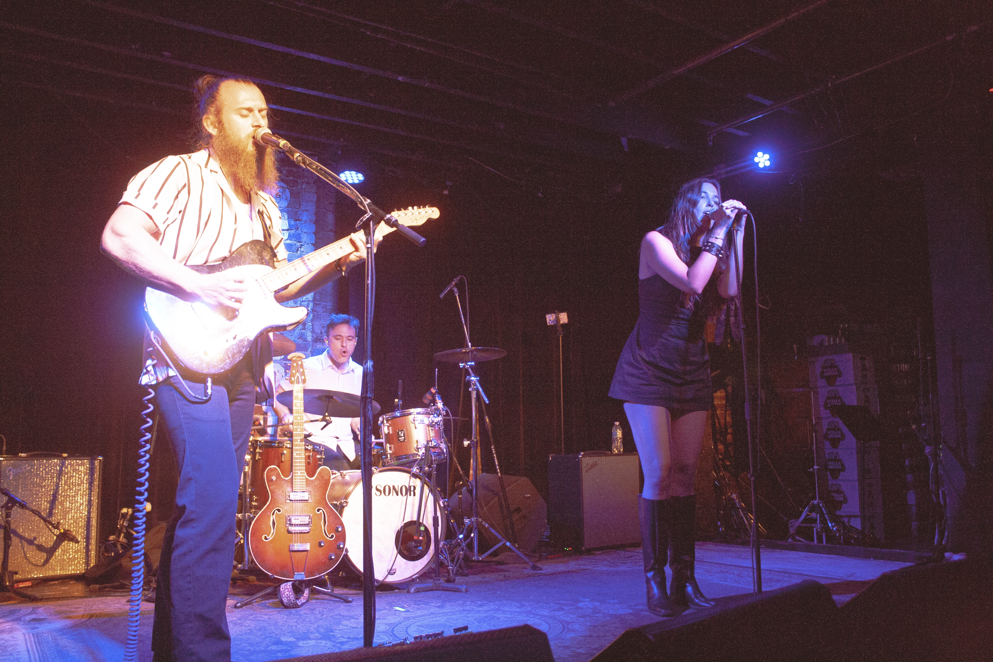 A band performing live on stage with a male guitarist and singer, and a female vocalist, under stage lights. The drummer is in the background, and there are guitars and amplifiers on stage.