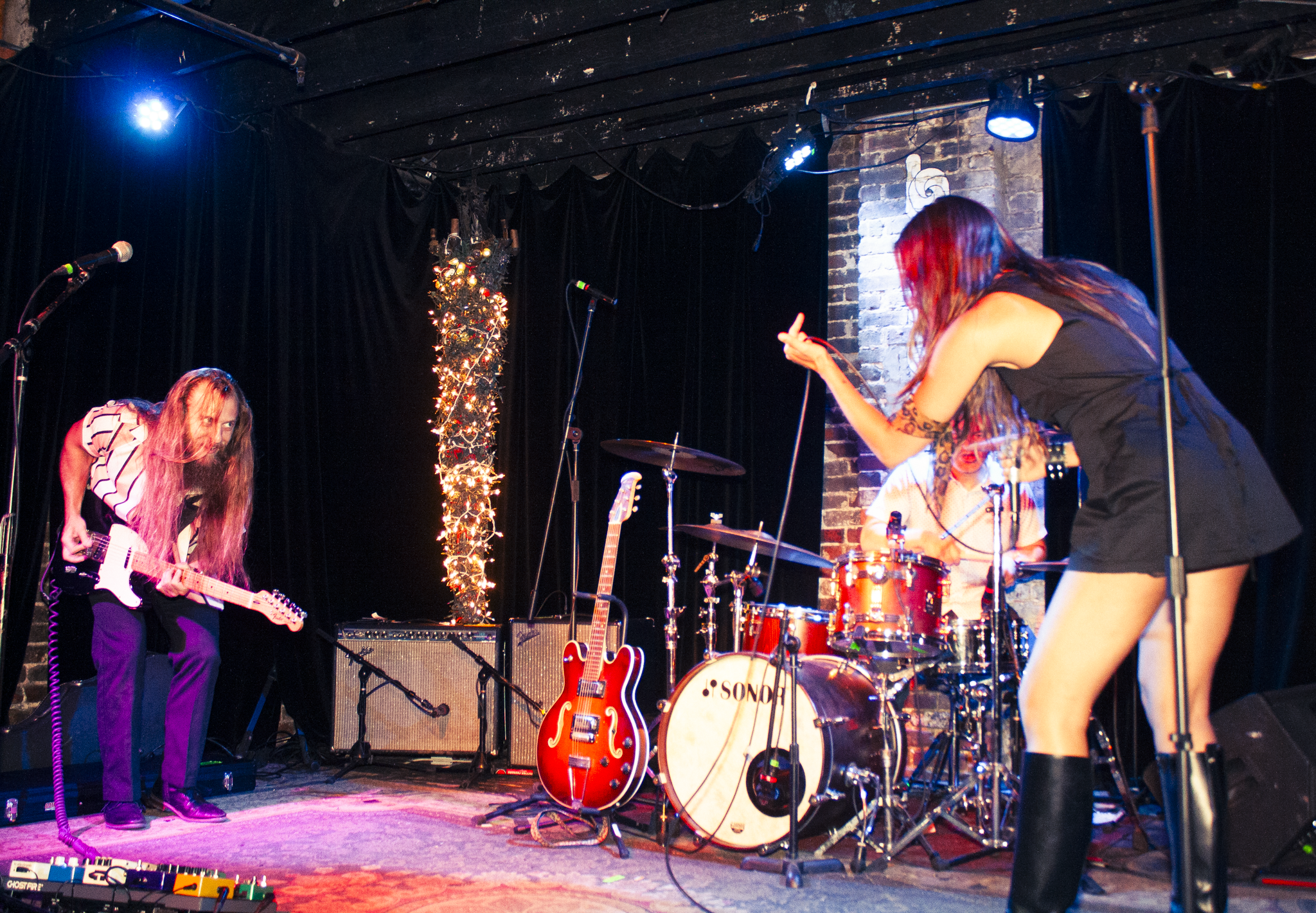 A live band performing on stage with musical instruments, including guitars, drums, and keyboards. The stage is decorated with string lights and a Christmas tree, with a brick wall as the backdrop. Two female musicians are visible, one with long red 