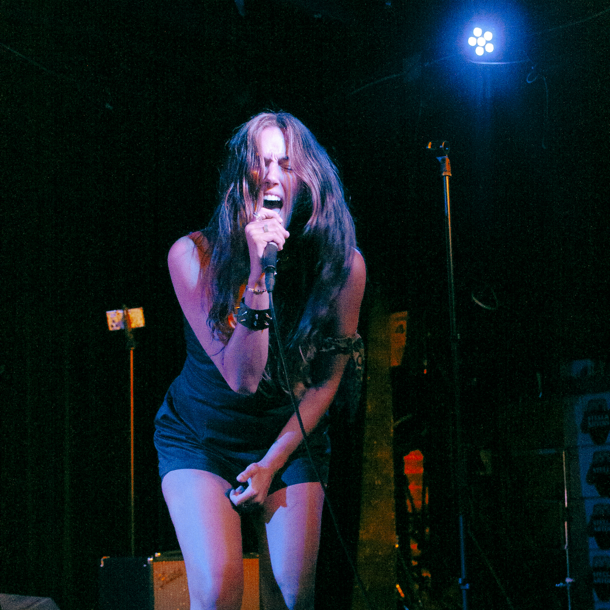 A woman with long, wavy hair singing passionately into a microphone on a dark stage, illuminated by blue and purple stage lights.