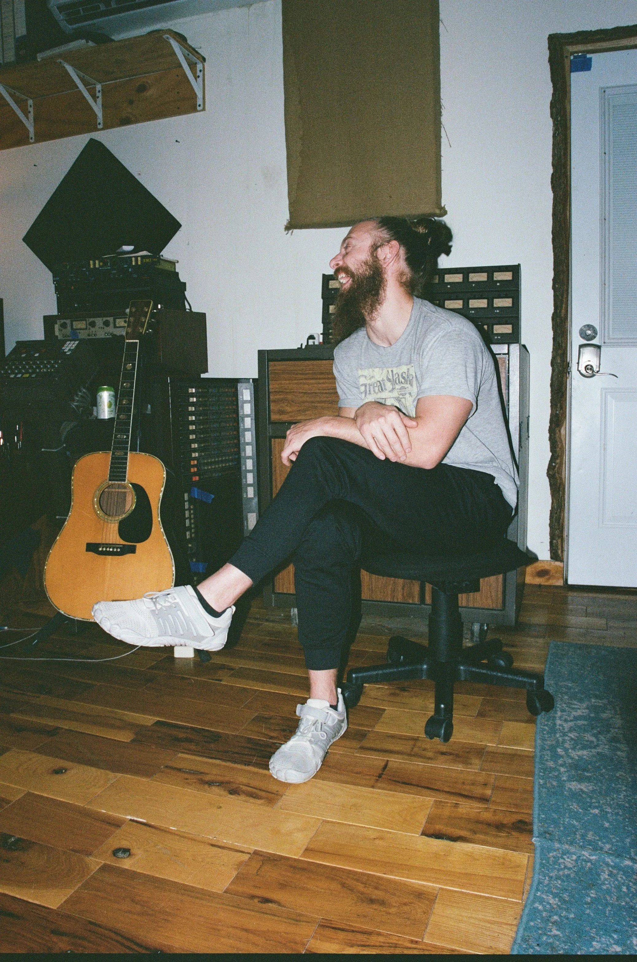 A man with a beard and long hair tied in a bun is sitting on a rolling office chair, smiling and laughing, in a music or recording studio with musical equipment and a guitar. Music being recorded at The Bomb Shelter Studios.