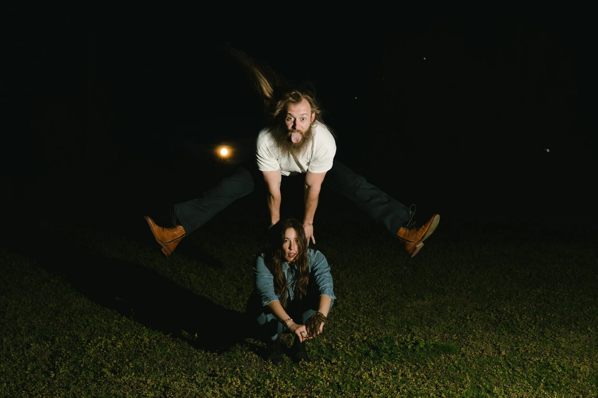 A man with long hair and a beard leans over a woman lying on the grass at night. The man is mid-jump with legs wide apart and tongue sticking out. The woman has long brown hair, wears a blue jacket, and looks up at the camera. The background is dark 