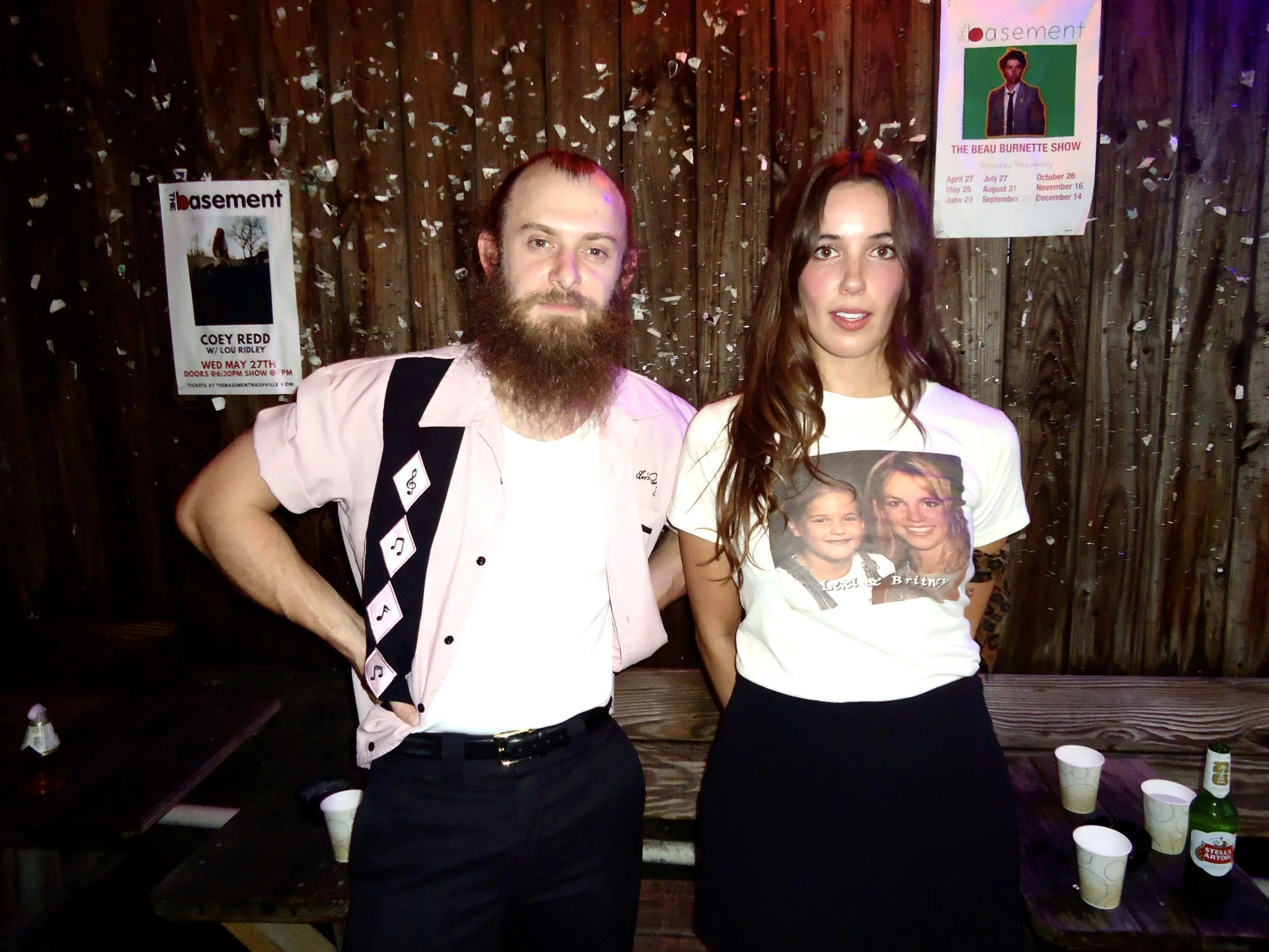 Two people standing in front of a wooden wall with posters, confetti falling around them, and cups on a table in front of them at an indoor party. The person on the left has a beard and styled hair, wearing a light pink shirt with musical notes on it