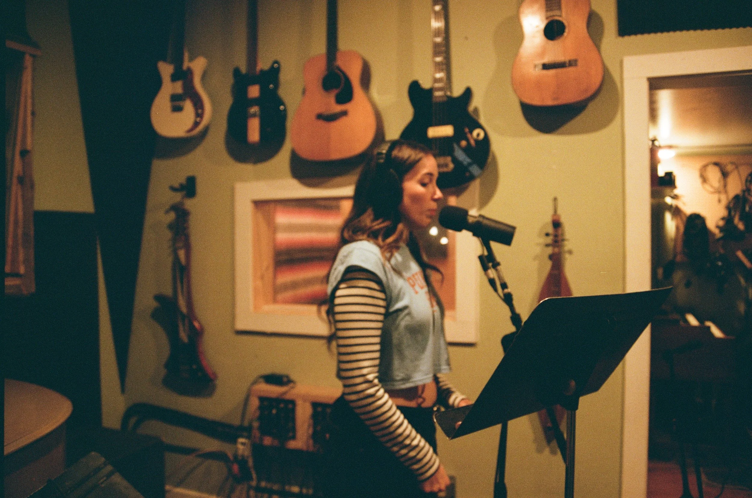 A woman wearing a striped shirt and vest is singing or speaking into a microphone in a music studio, with guitars hanging on the green wall behind her. Music being recorded at The Bomb Shelter Studios.