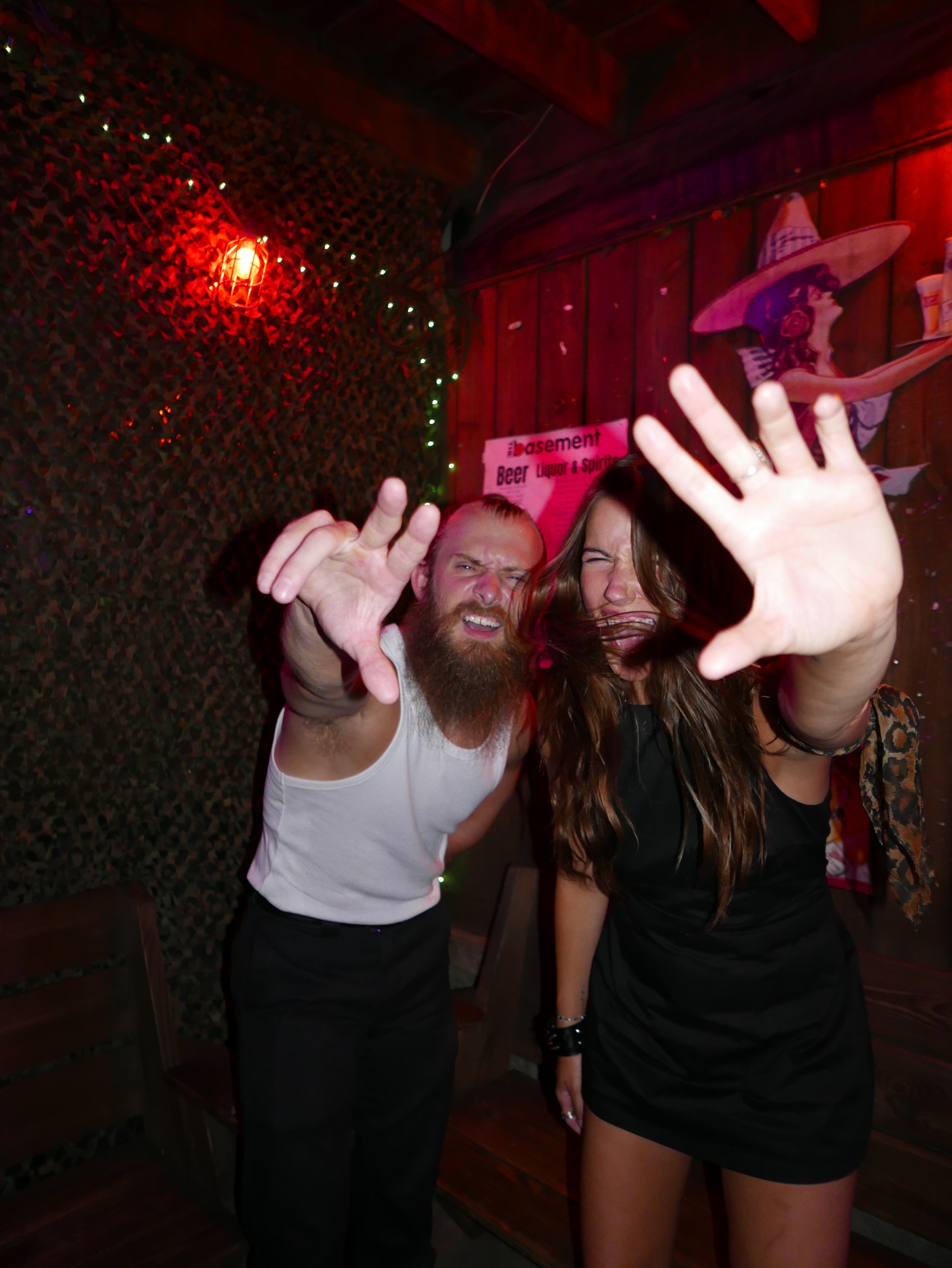 Two people are dancing closely in a dimly lit bar with red lighting, a sign in the background reads "basement Beer Liquor & Spirits," and there is a wall decorated with string lights and a silhouette mural of a woman with a sombrero.