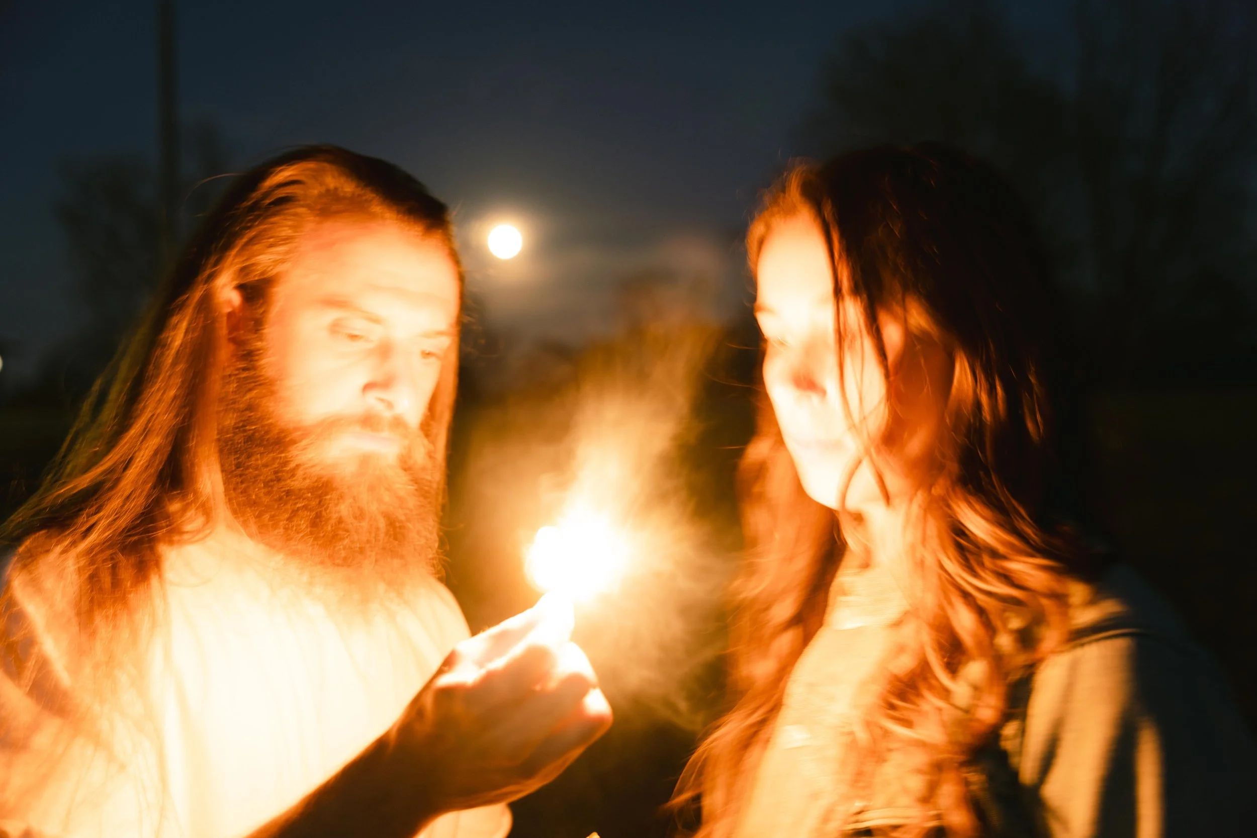 Two young women with long hair holding a lit sparkler at night, with the moon in the background.