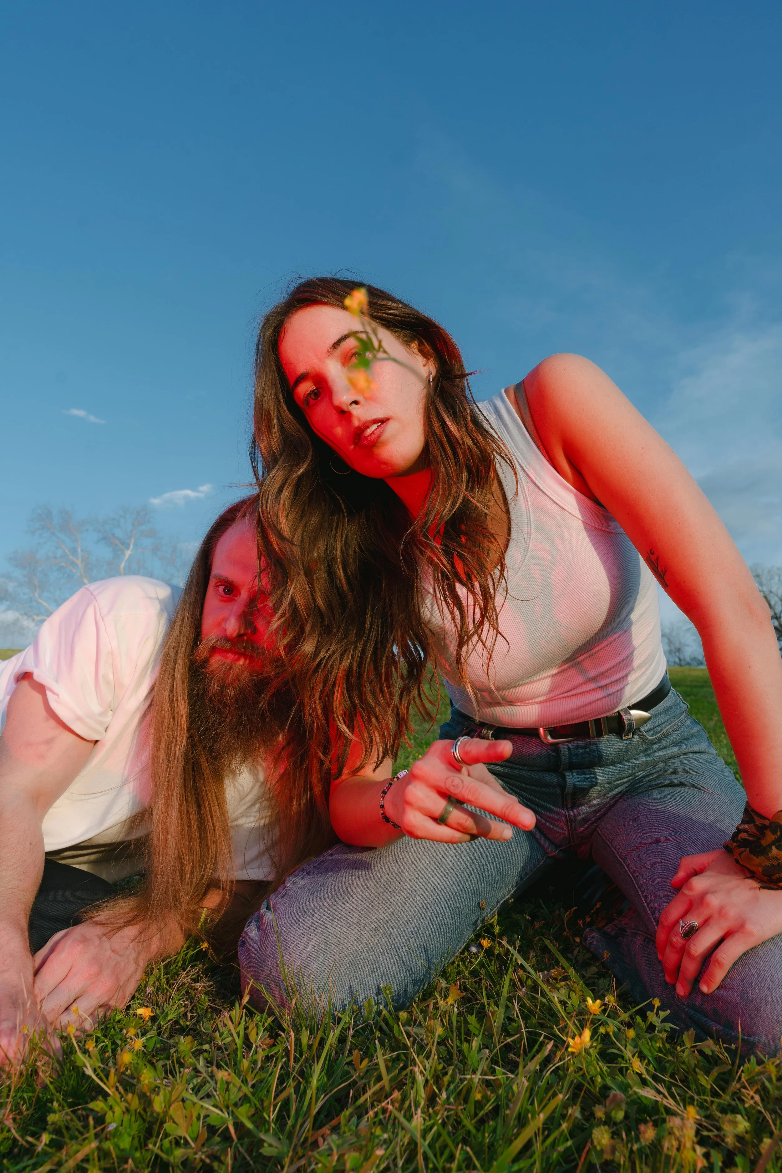 A young woman with wavy brown hair making a peace sign gestures with her hand, sitting on grass outdoors, with a man with long hair and a beard lying next to her.
