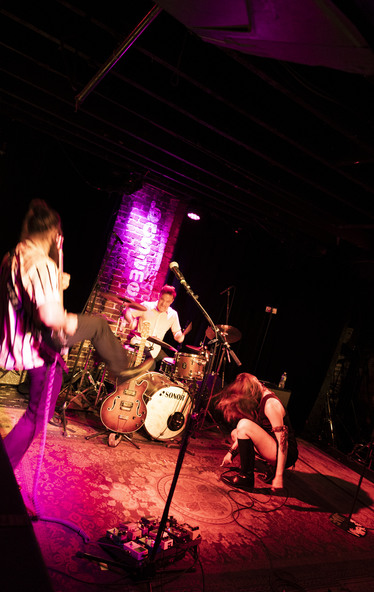 A band performing on stage with purple lighting, featuring a drummer, a guitarist, and a female singer in black boots and a black outfit, with music equipment and pedal effects visible, in a venue with brick walls and a sign that reads 'Regency'.