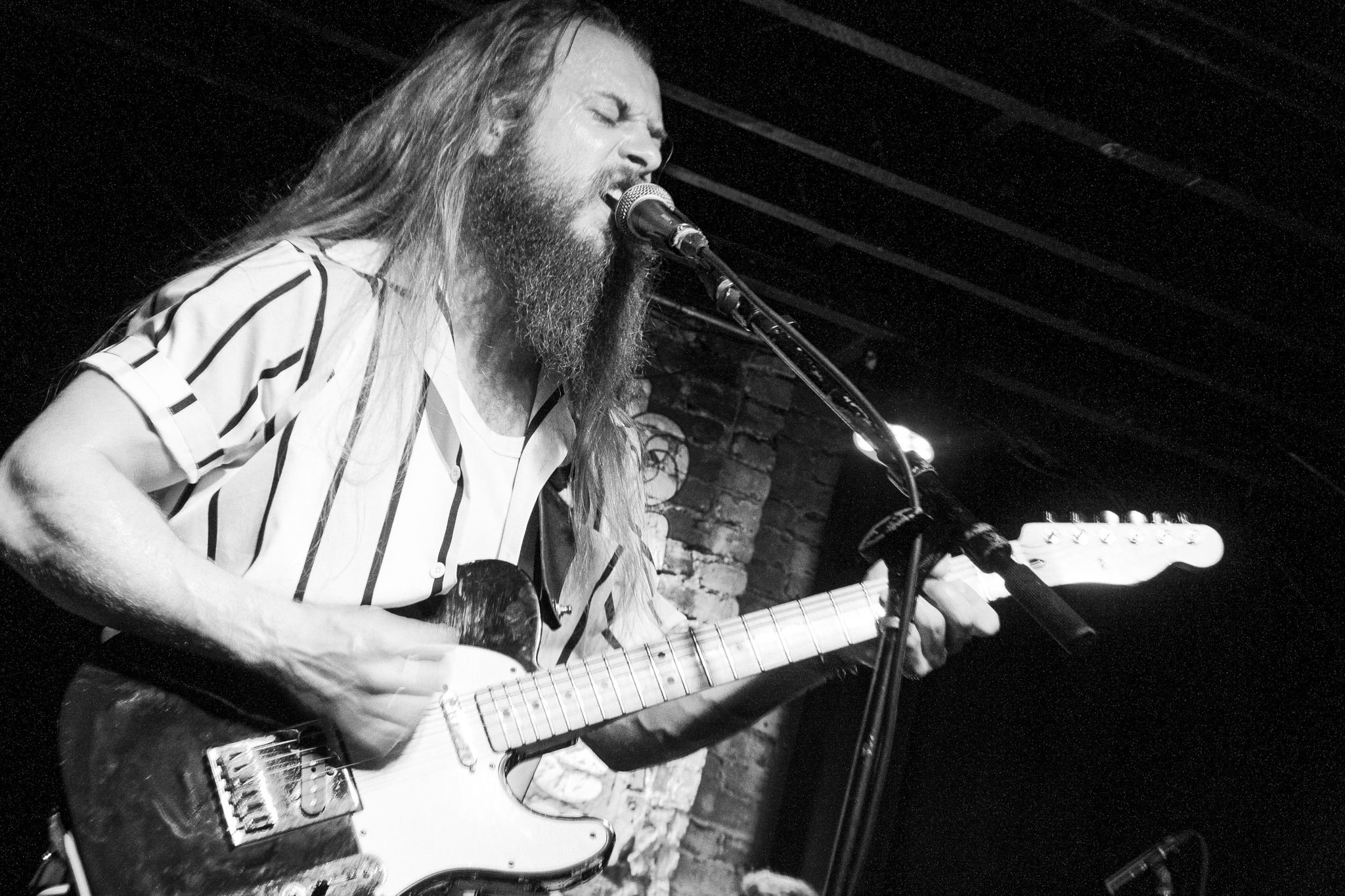 A male musician with long hair and a beard passionately singing into a microphone while playing an electric guitar, on stage with a brick wall background.