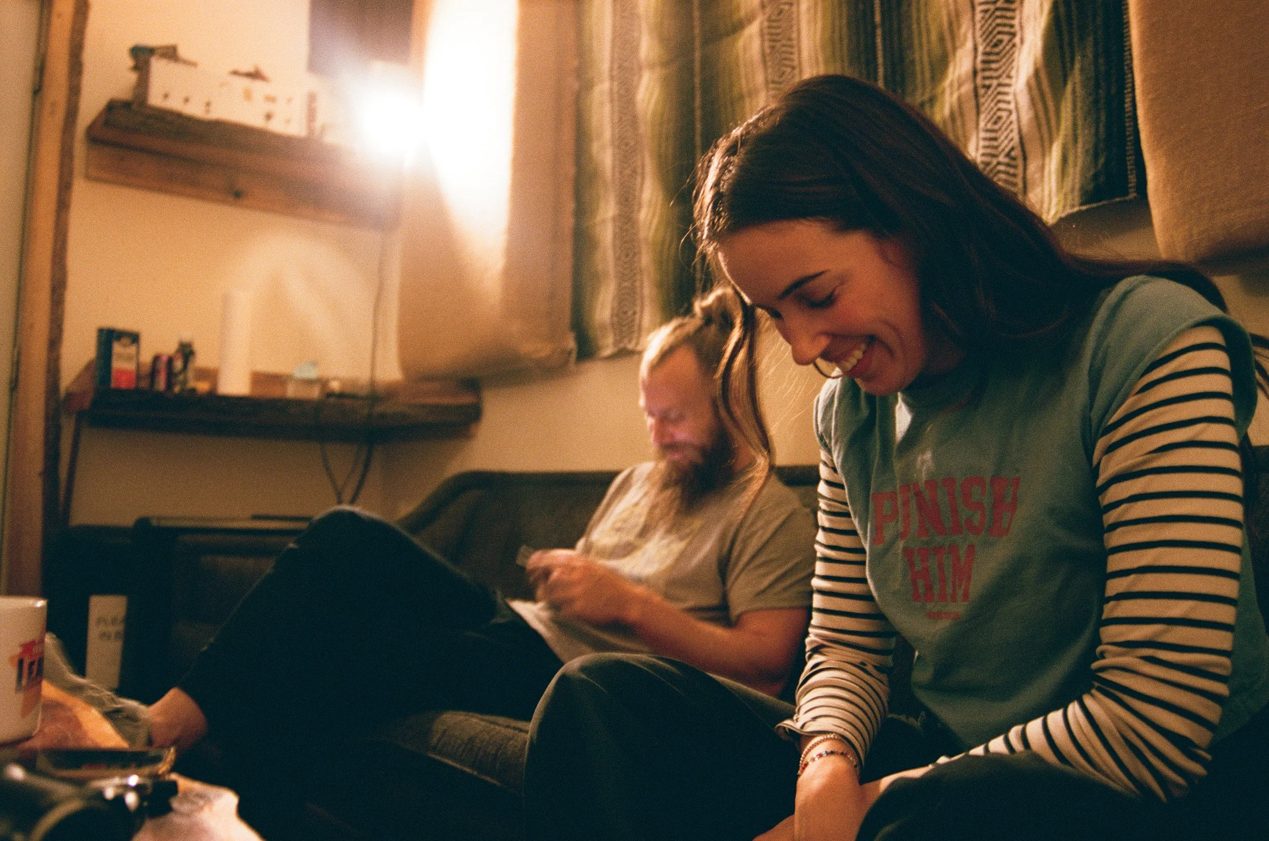 Young woman with long dark hair wearing a striped shirt and smiling while sitting on a couch. Behind her, another person with long hair appears to be looking at their phone. Music being recorded at The Bomb Shelter Studios.