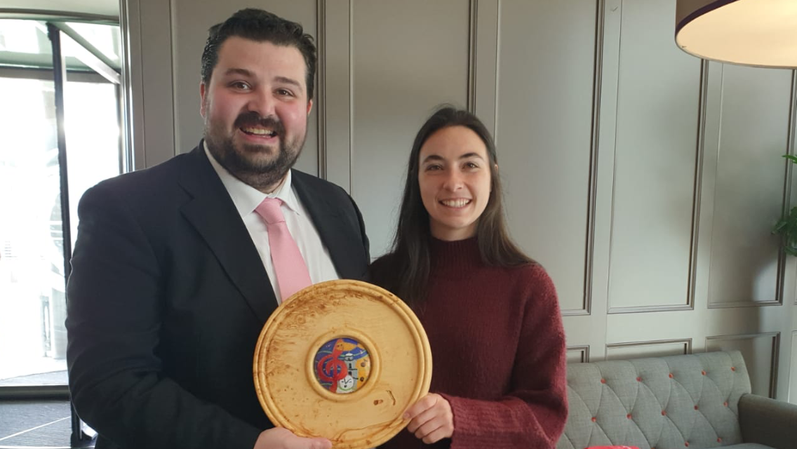 A man in a dark suit and pink tie holding a wooden plaque with a colorful ceramic center, standing next to a woman in a maroon sweater, both smiling indoors.