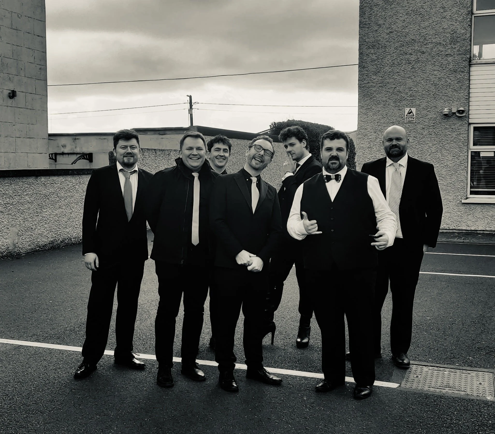 A group of seven men dressed in formal attire, standing outside near a building, with cloudy sky overhead.