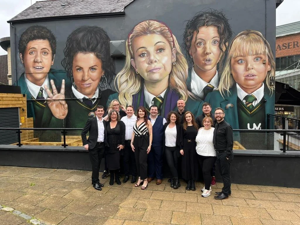 A group of ten people standing in front of a large mural of characters from the TV show 'Stranger Things', which depicts five young characters with distinctive hairstyles and expressions. The group is smiling and dressed in semi-formal attire.