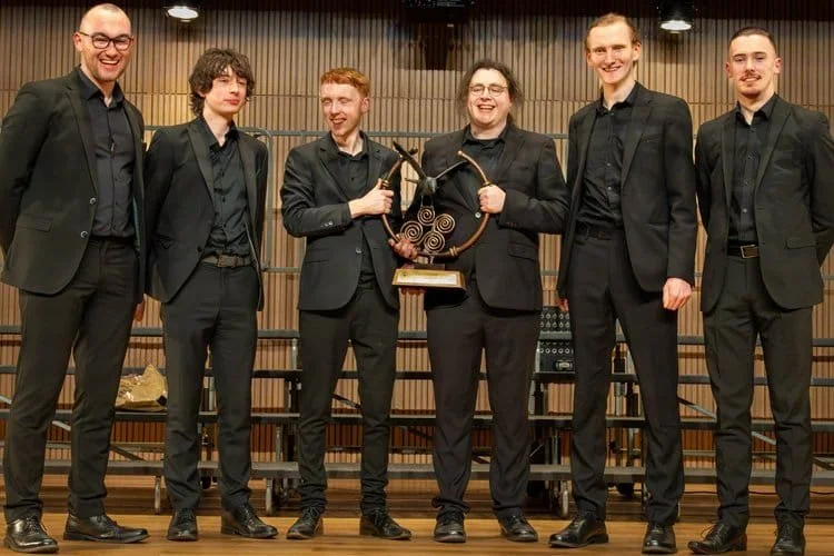 Six people dressed in black suits standing on a stage, holding a spiral-shaped trophy, with wooden paneling and stage equipment behind them.
