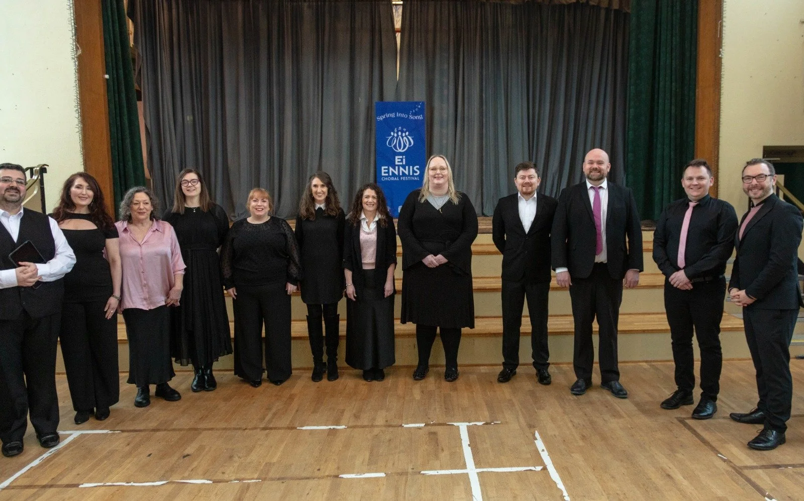 A group of 13 people standing on a stage in front of a blue banner that reads 'Spring Into Song. Ei ENNIS Choral Festival.' They are dressed in formal black and white attire, smiling for a group photo indoors with wooden flooring and a dark curtain b