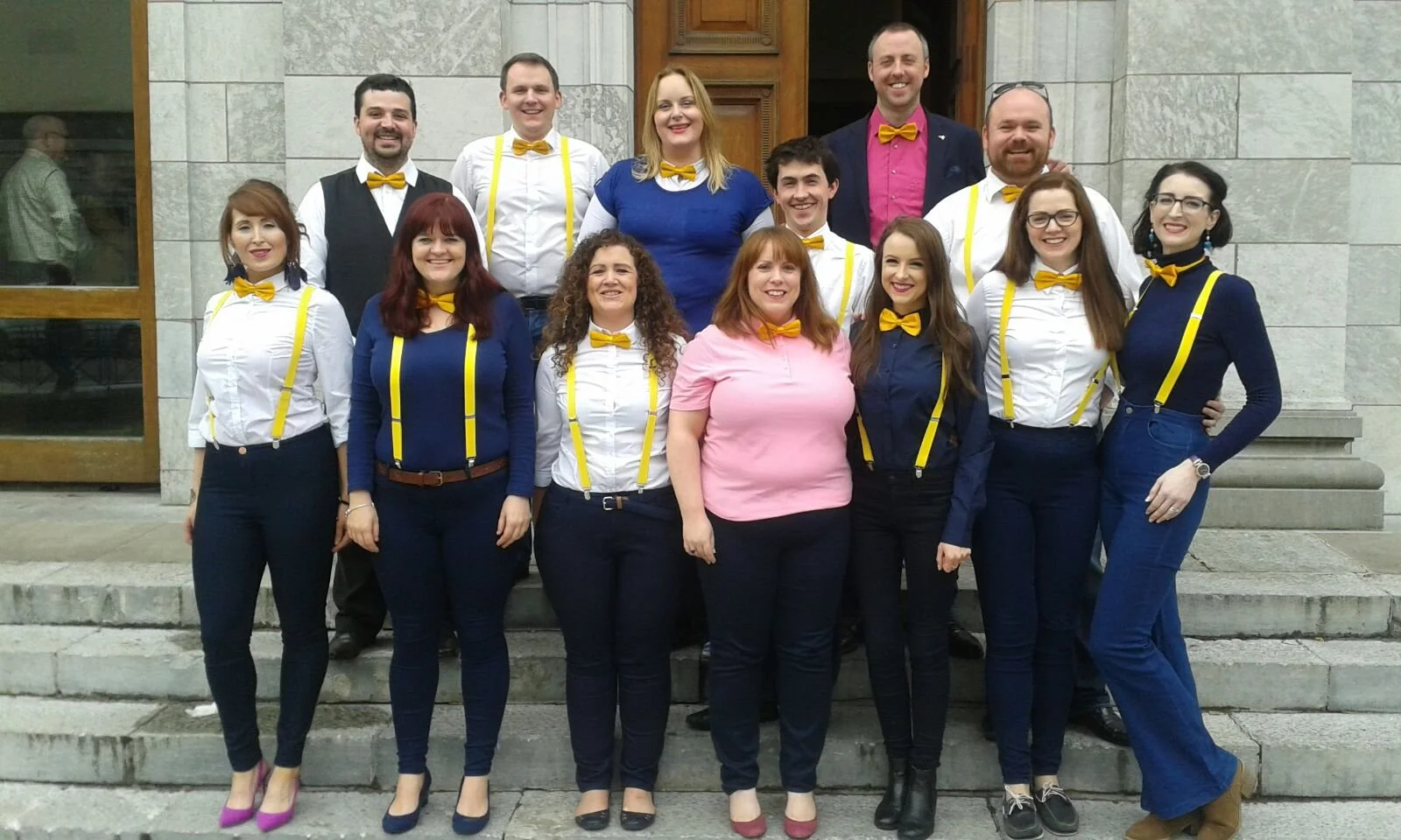 Group of people standing on steps outside a building, dressed in colorful clothing with yellow suspenders and bow ties, posing for a photo.