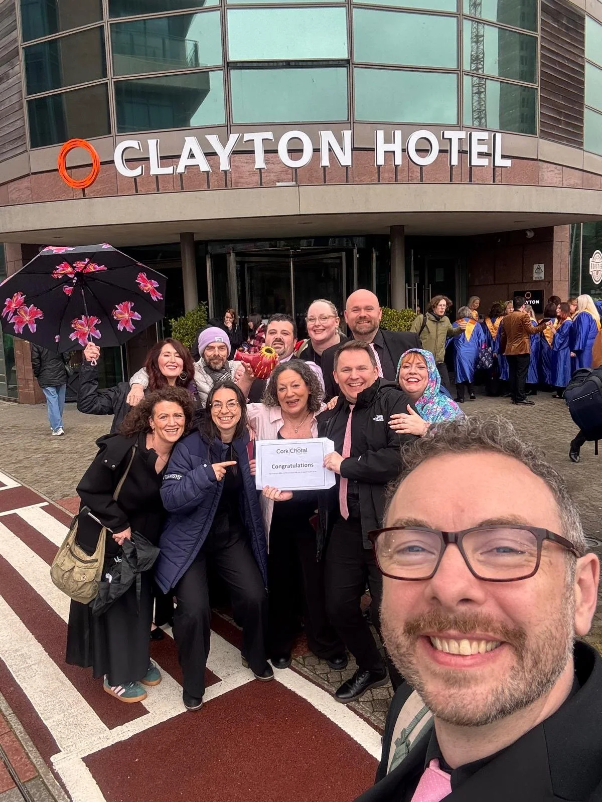Group of people smiling and celebrating outside the Clayton Hotel, with some holding a sign that says 'Cork Choral Congratulations', and a woman holding a pink and yellow object, with a person in the background holding a pink floral umbrella, and others wearing graduation gowns.