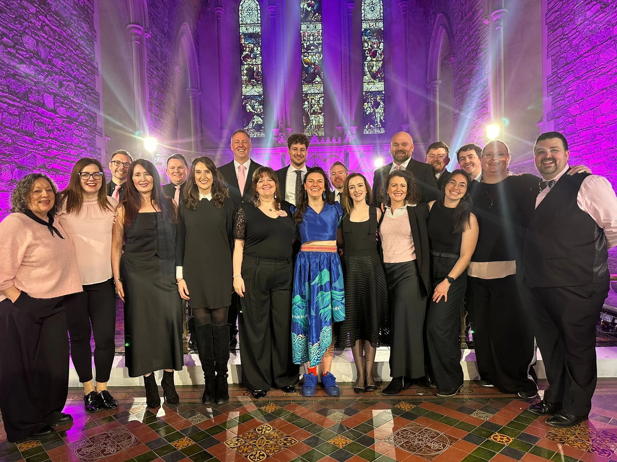 A group of people dressed in formal attire standing together inside a church with stained glass windows and purple lighting.