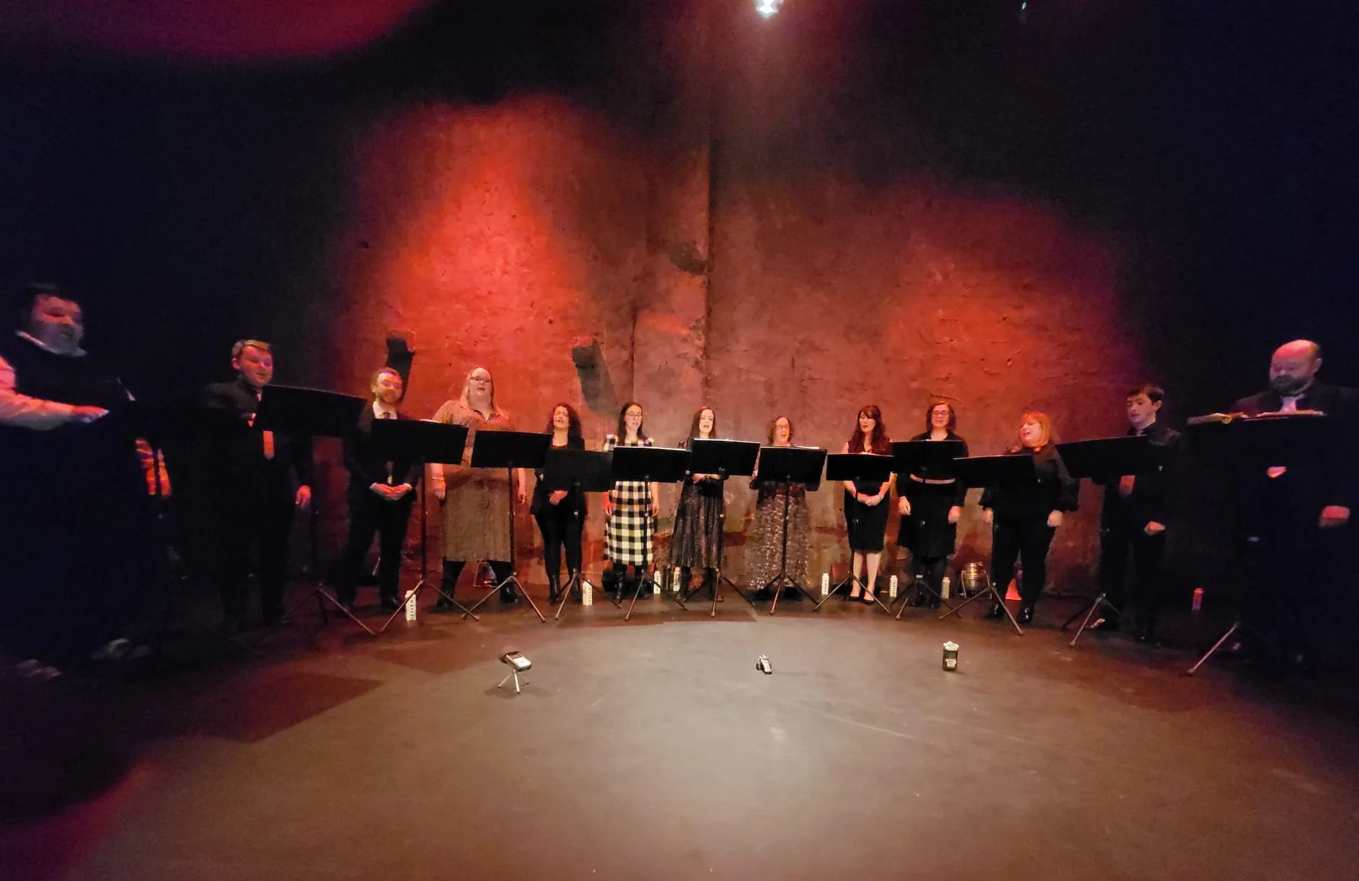 A group of people standing on a stage in a theater, possibly during a performance or ceremony. There are microphones and music stands in front of them, and the background features a large brick wall illuminated with red and orange lighting.