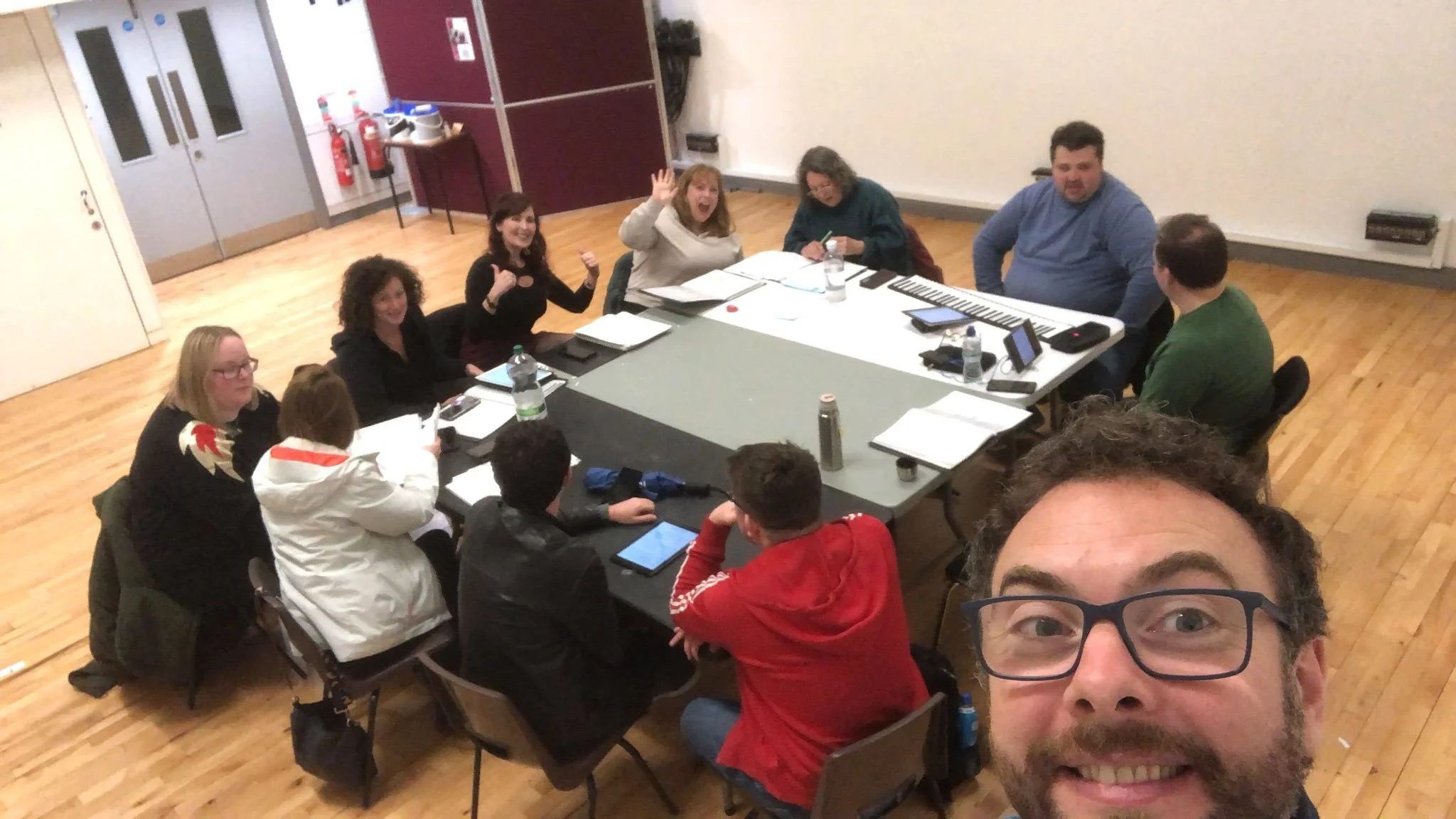A group of people sitting around a table in a meeting or workshop, with some smiling and waving at the camera. There are notebooks, water bottles, and electronic devices on the table in a room with wooden floors and plain walls.