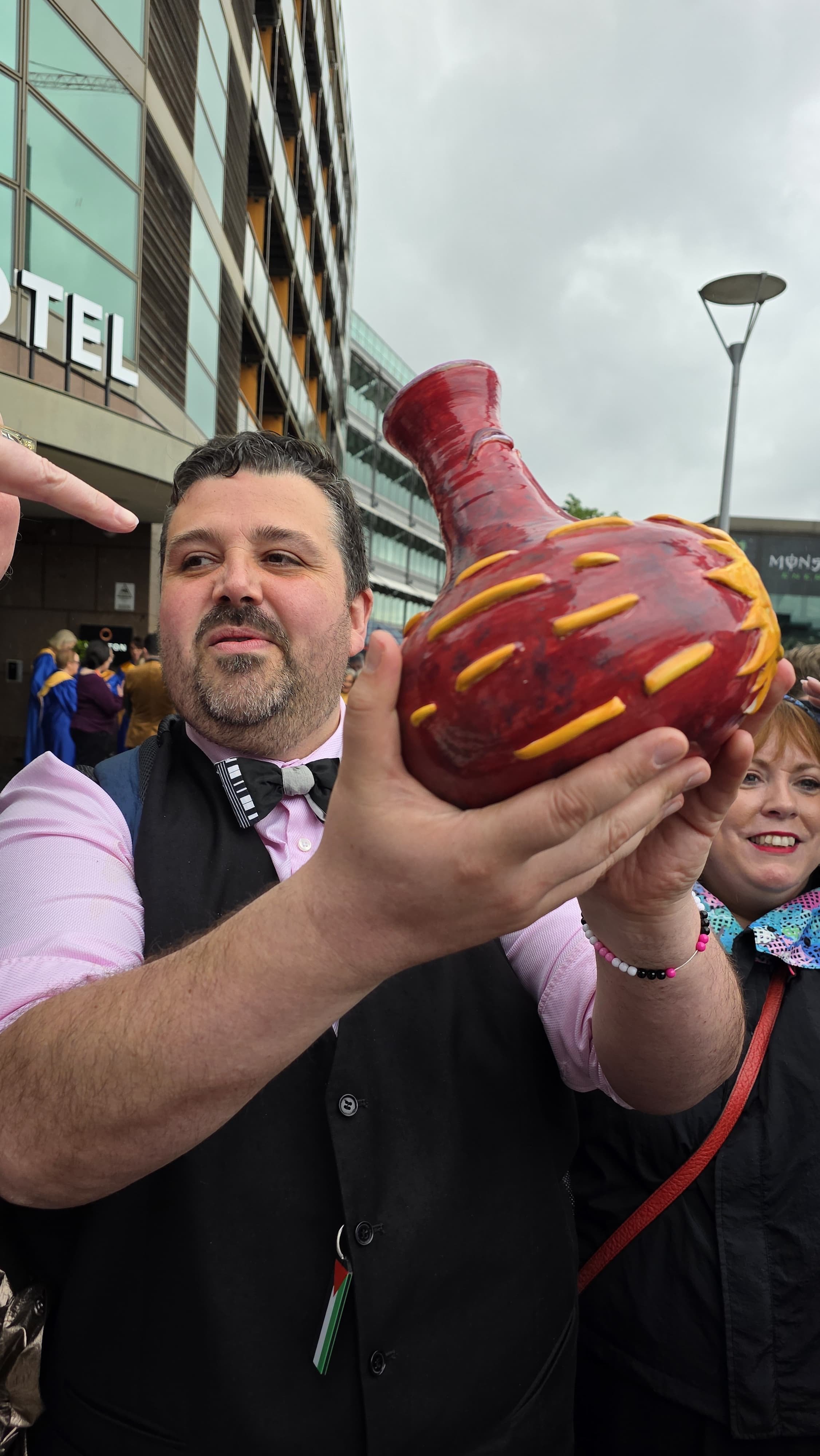 A man in a black vest, pink shirt, and bow tie holding a large, decorative red and yellow ceramic vessel in an outdoor setting with other people and a modern building in the background.