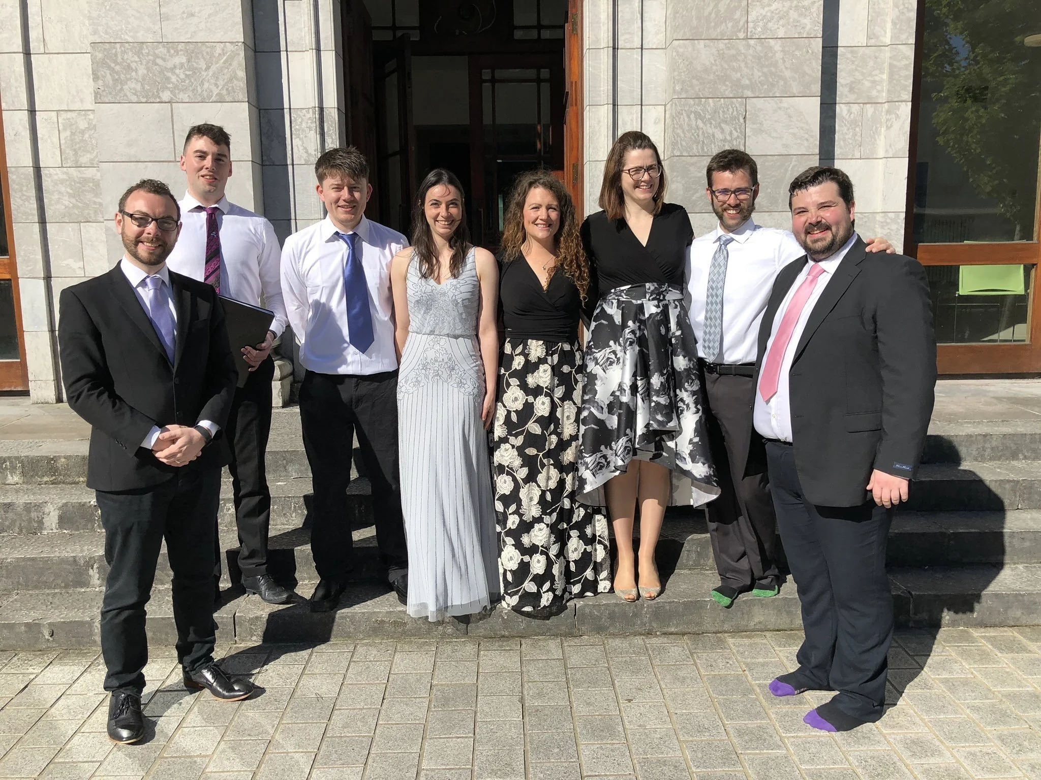 Group of eight people dressed in formal and semi-formal attire standing on steps outside a building, smiling for a photo.