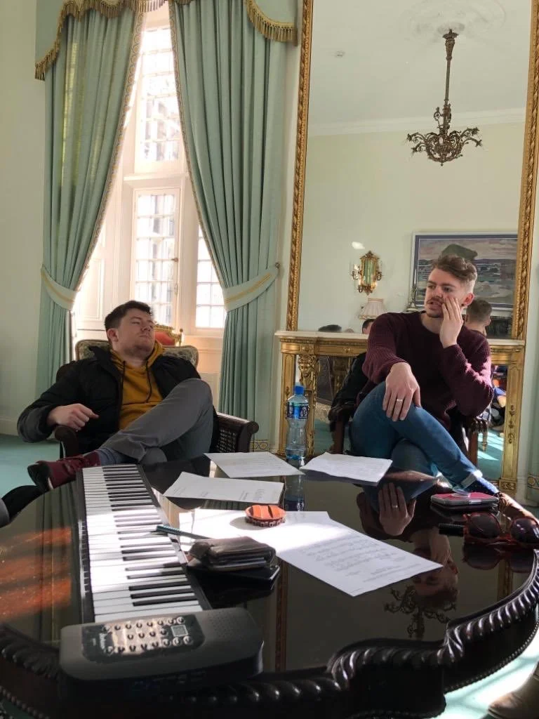Two men sitting at a large, ornate table with papers, a water bottle, a remote, and a keyboard on it. One man is seated in a chair with his legs crossed, the other is resting his head on his hand and looking away. The room has green curtains, a large