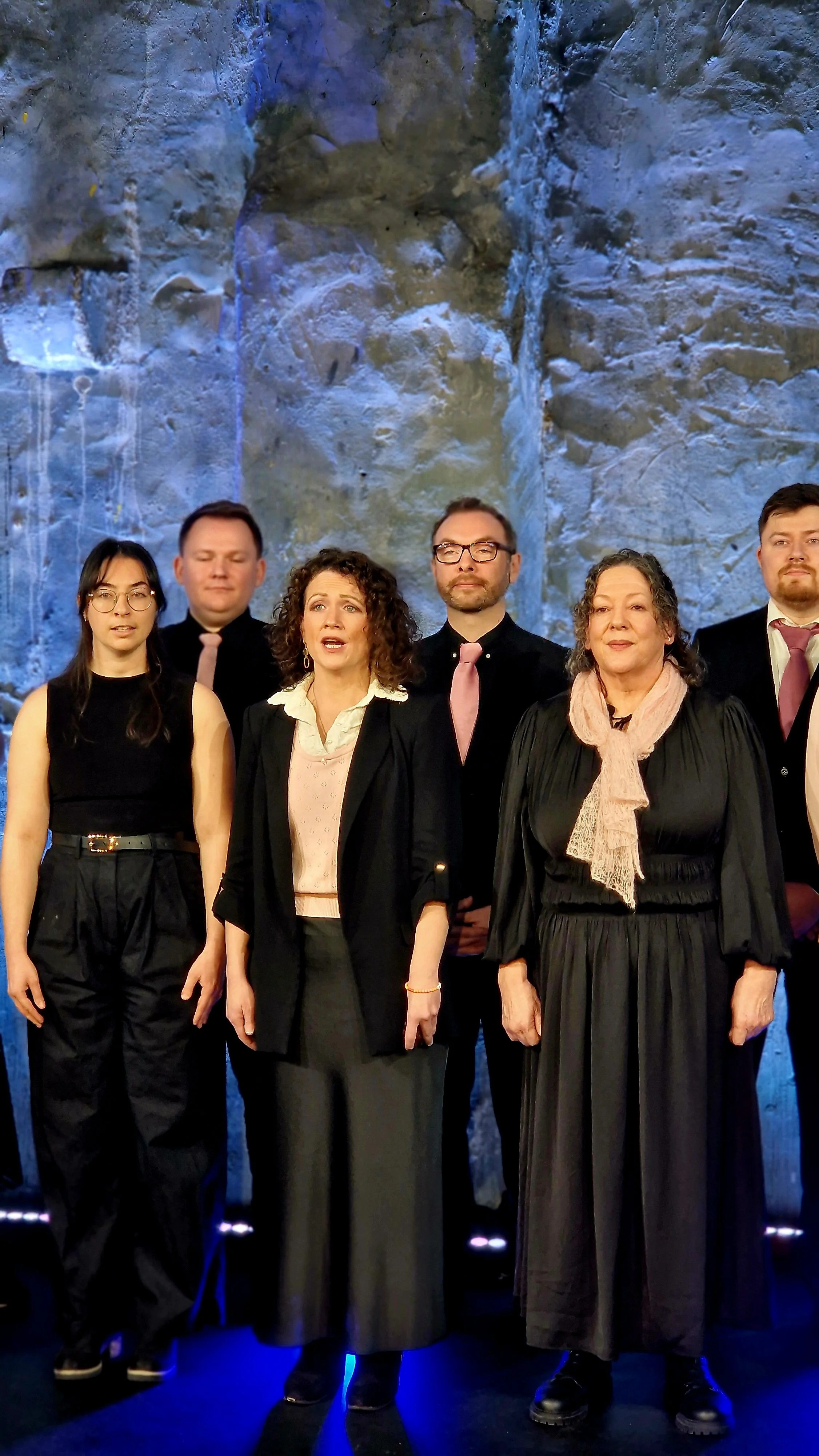 Group of six people standing in front of a textured blue-gray wall, dressed in formal and semi-formal attire, with serious and attentive expressions.