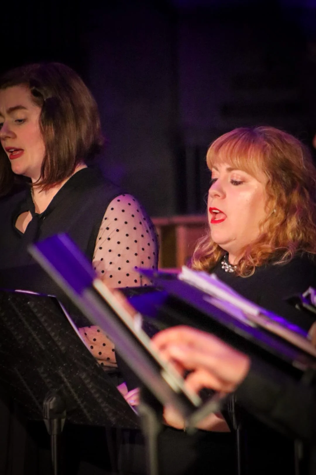 Two women singing or performing, one with dark hair and the other with red hair, in a dimly lit setting with music stands and sheet music.