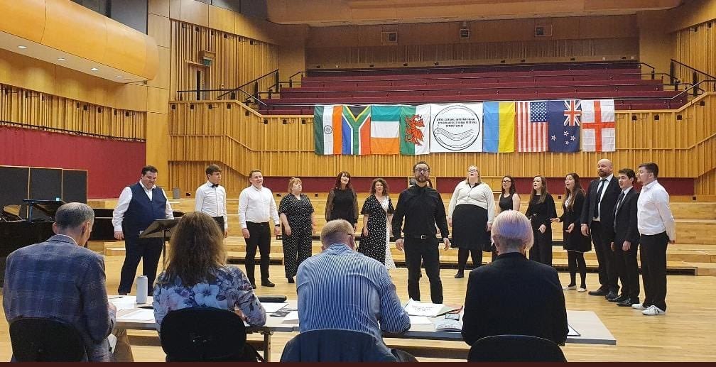 A choir of 12 young adults performing in a music rehearsal space, with four judges seated in front, and flags from South Africa, India, Ireland, Wales, United States, and England hanging on the wall.