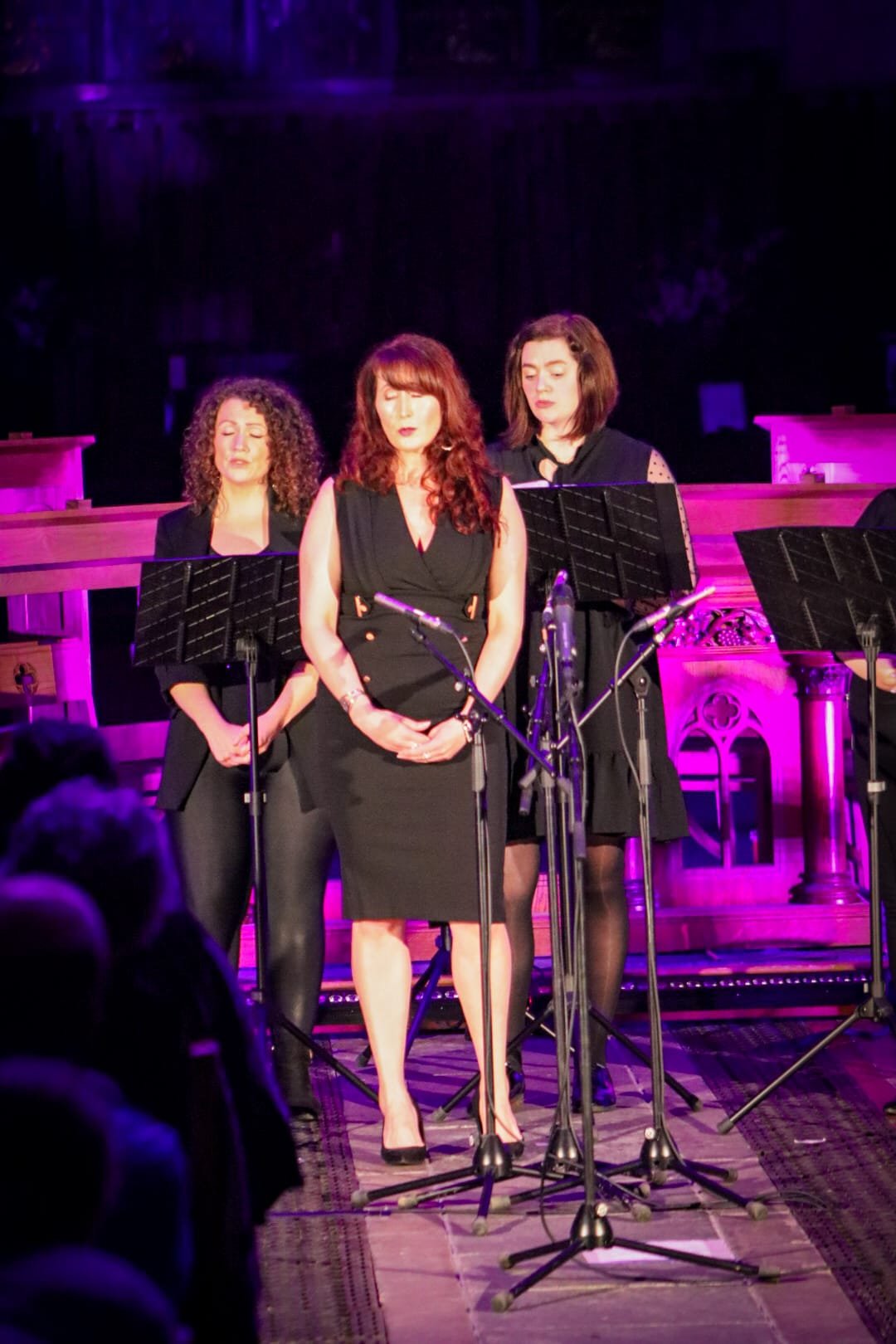Three women standing on stage with music stands, in front of a purple-lit background, wearing black attire, with microphones set up in front of them, in what appears to be a musical performance or concert.