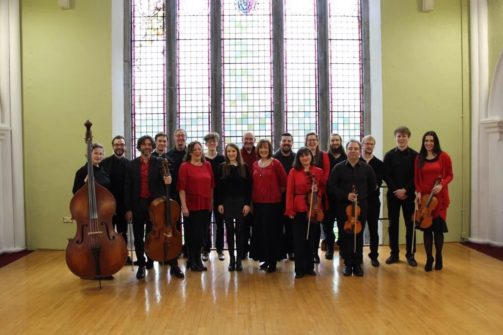 A group of 16 musicians, including four holding string instruments, standing indoors in front of a large, colorful stained glass window, with light green walls and a wooden floor.