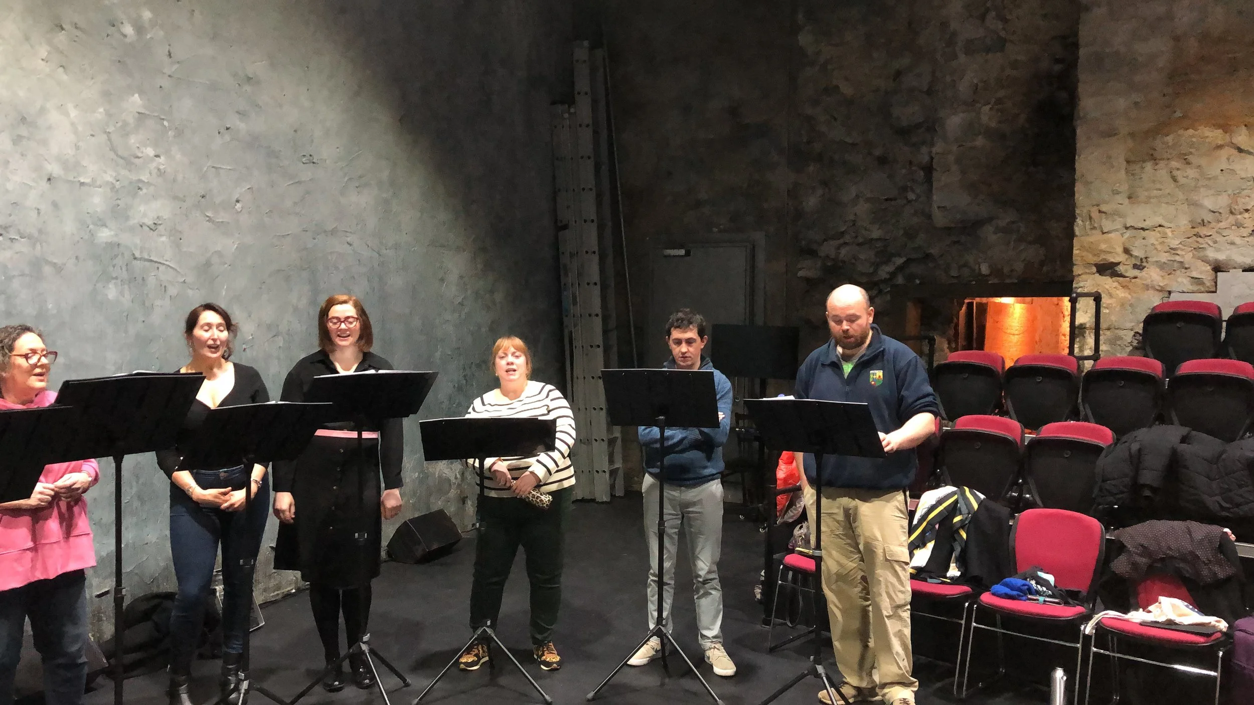 Group of people standing and singing in a rehearsal space with black music stands, red chairs, and exposed stone and concrete walls.