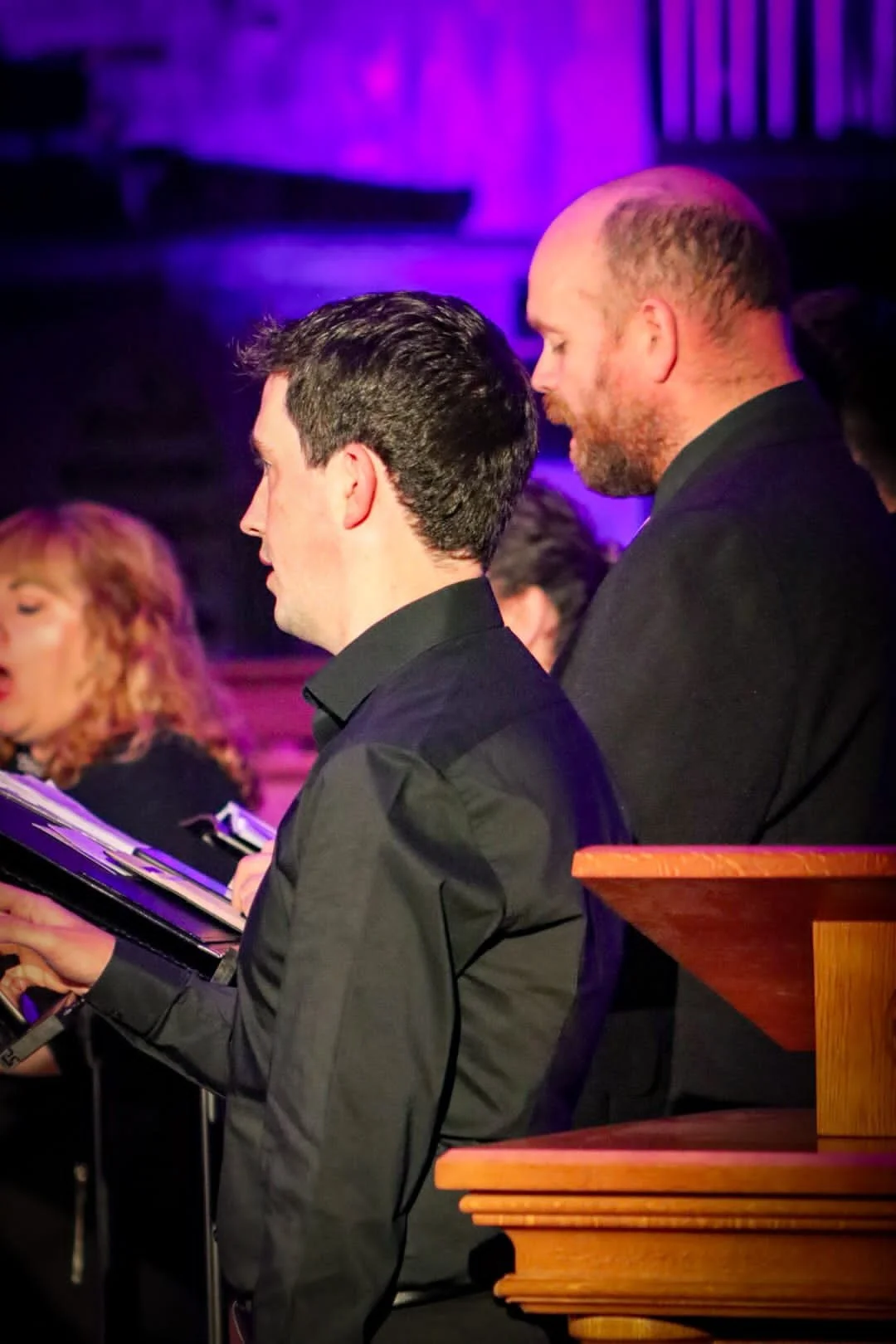 Two men and a woman sitting at a concert or event, with purple lighting in the background, and one man reading from a music score.