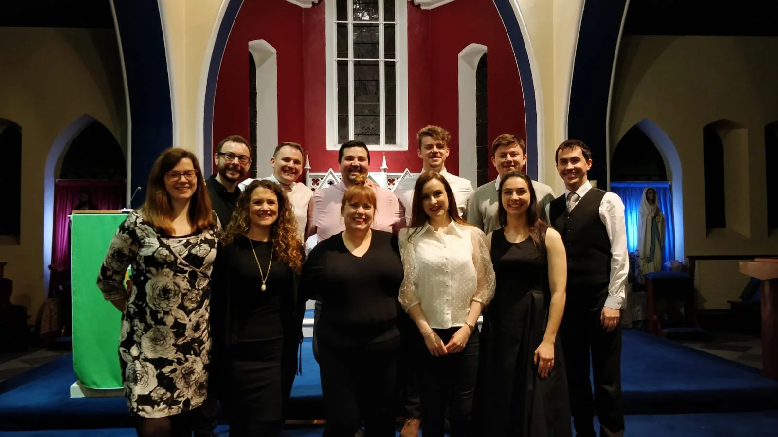 Group of eleven people, men and women, dressed in semi-formal attire, standing together inside a church with stained glass windows and religious statues in the background.