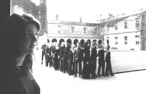 A woman with long hair looking to her right in the foreground, with a line of people, mostly young men, standing in a line outside a historic building with arches and windows in the background.