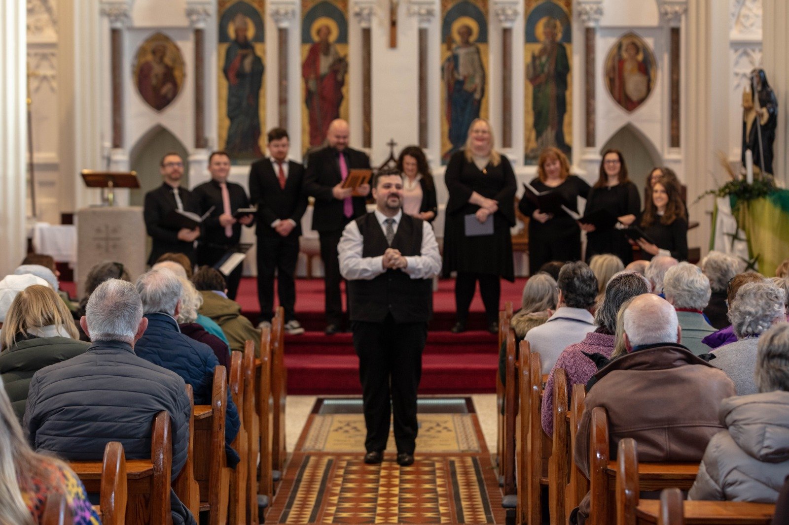 A choir performing in a church with a congregation watching, led by a man standing in front, with the choir standing on the steps of the altar behind him.