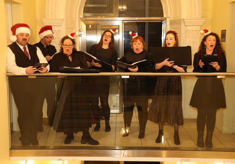 Six people singing Christmas carols on a balcony, wearing Santa hats, holding songbooks or sheet music, in a festive setting.