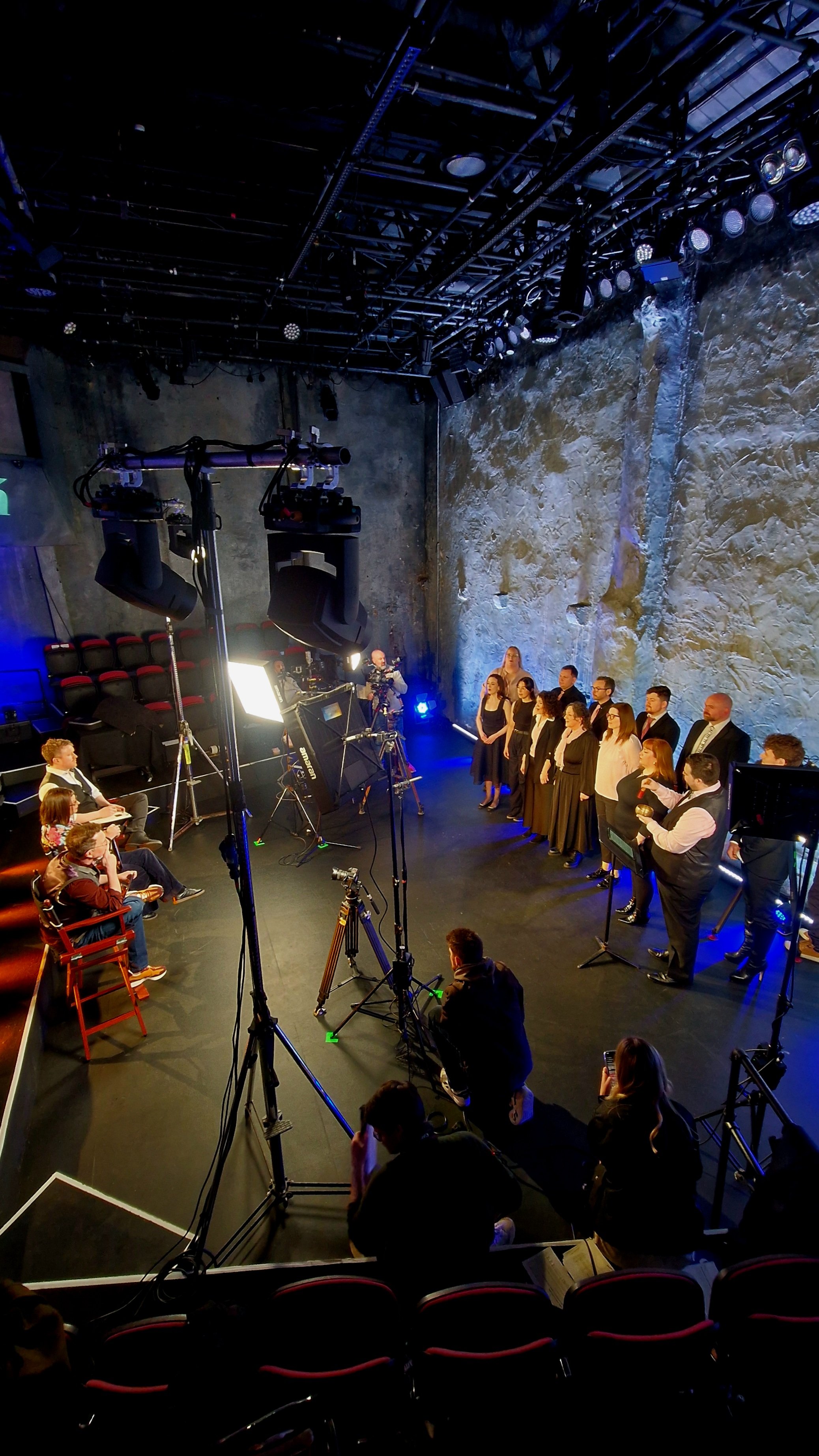 People on stage during a performance or event, with audience seated in front, in a room with stone walls and professional lighting equipment overhead.