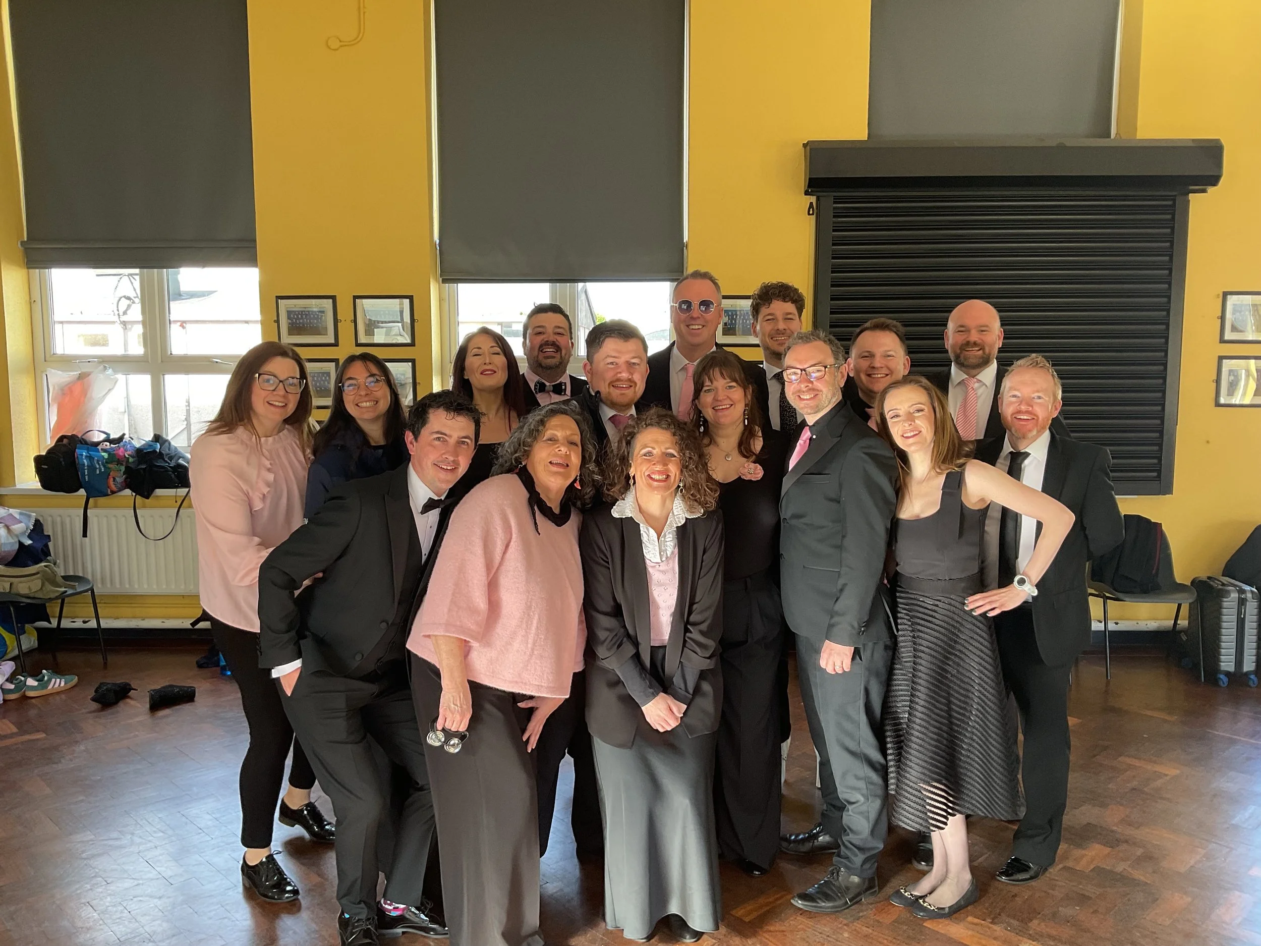 A group of 17 people dressed in formal and semi-formal attire smiling and posing together inside a yellow-walled room with large windows, some wearing tuxedos and evening dresses, celebrating at a social event or party.