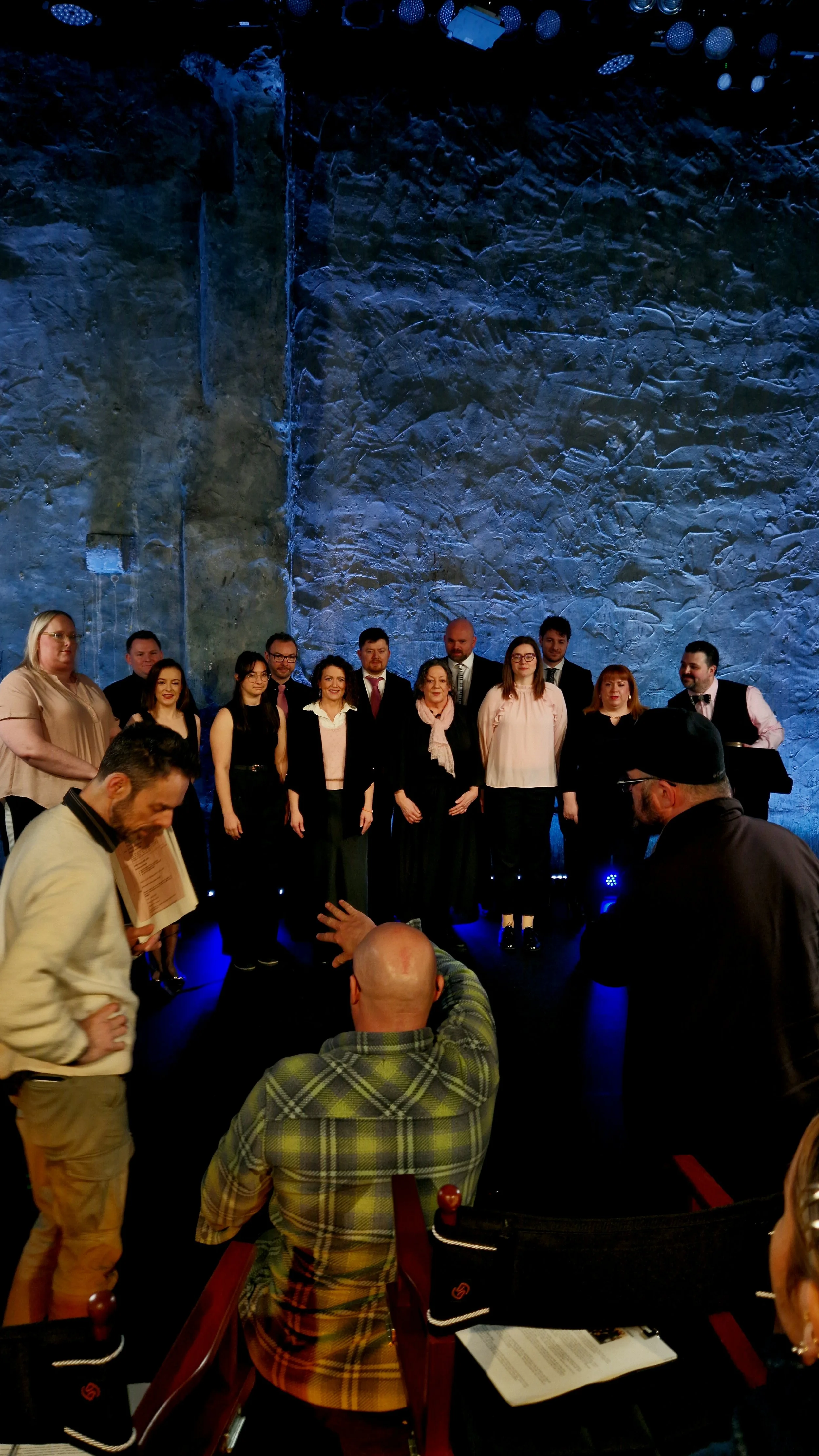 Group of people standing on stage during a formal event, with a blue textured wall in the background and audience members seated in front.