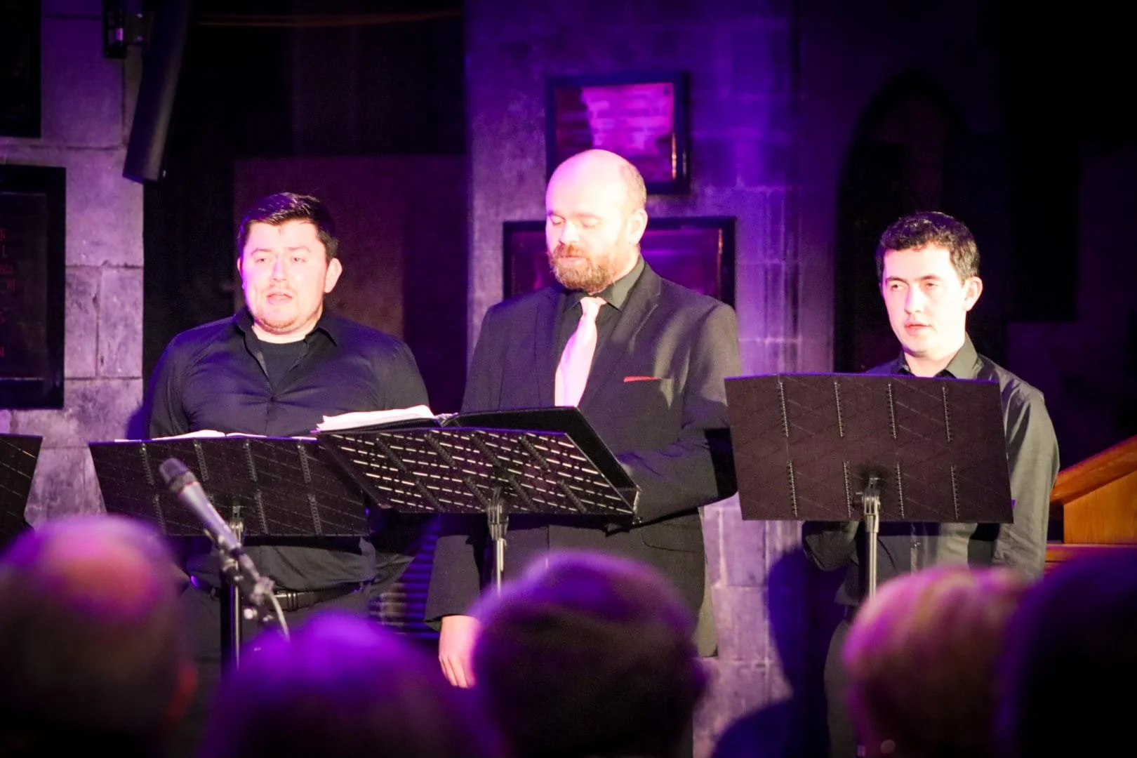 Three male singers performing on stage with music stands and microphones in front of them, in a dimly lit venue with brick walls.