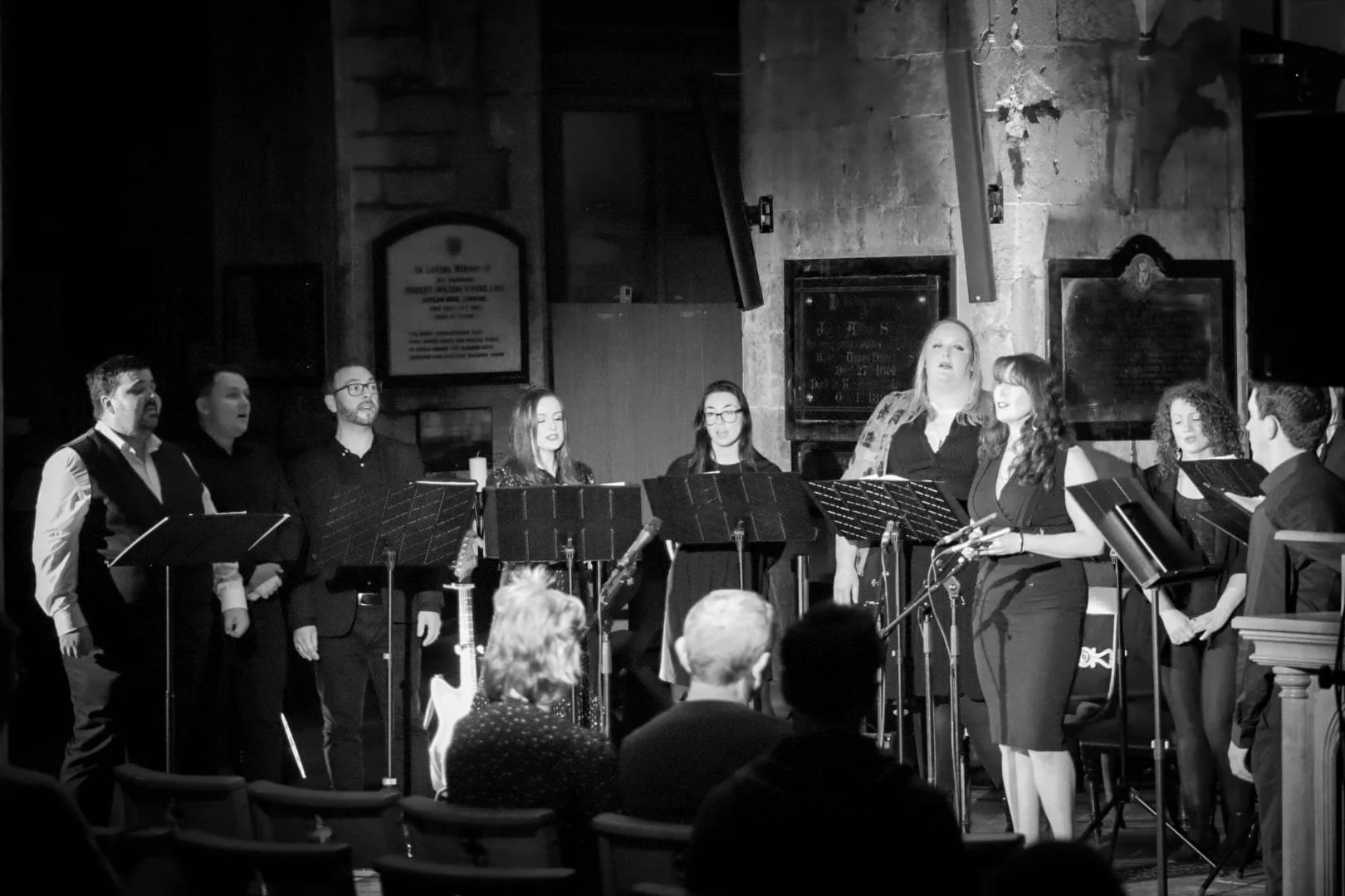 A choir or vocal group performing on stage inside a church or concert hall. The group includes men and women standing with music stands, with some holding microphones, and a woman in front singing. Audience members are seated in front of them.