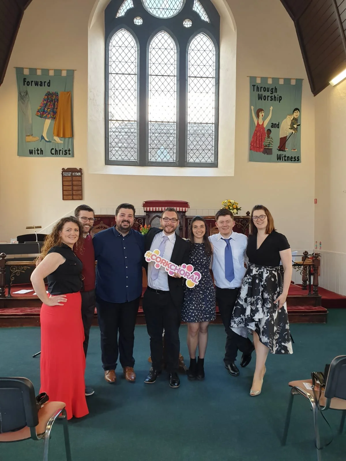 Group of seven people standing inside a church, smiling for a photo. The church has stained glass windows, banners on the walls with religious messages, and a choir sign. The group includes four women and three men, dressed in formal and semi-formal attire.