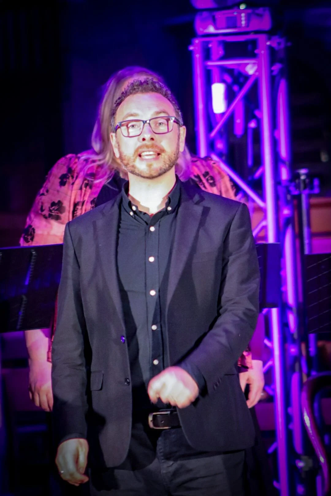 A man with glasses and a beard dressed in a black suit and black shirt standing in front of a purple-lit stage with a person in a floral shirt in the background.