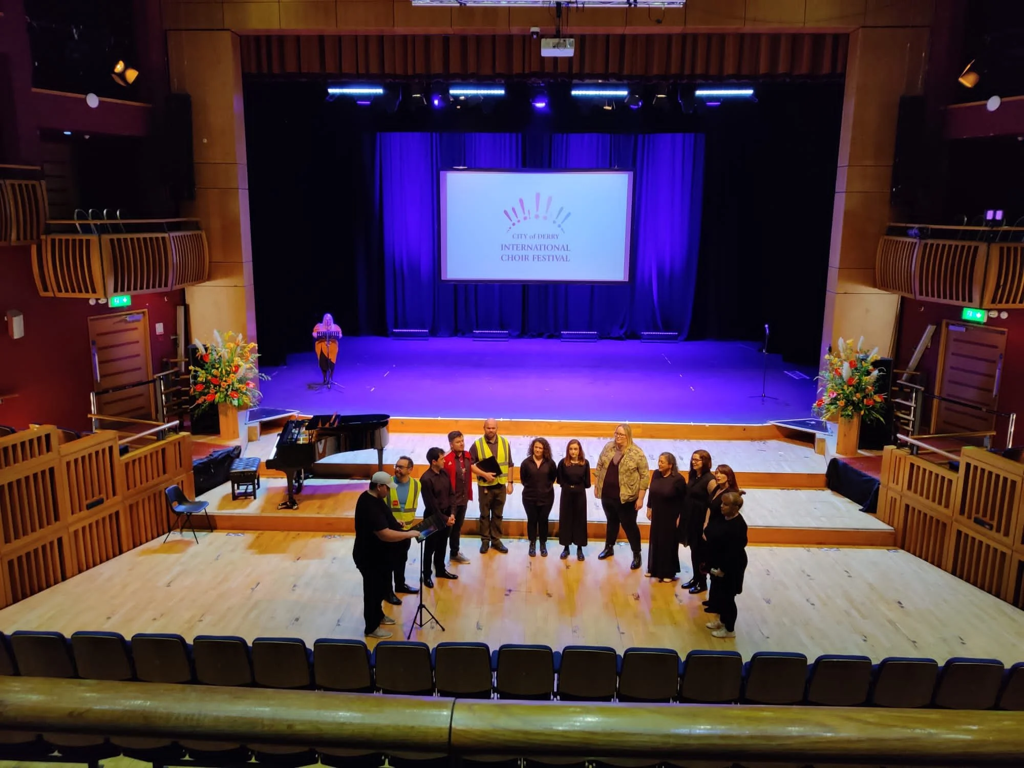 Stage inside a theatre with a large screen displaying the logo and text for the City of Derby International Choir Festival. People are gathered on the stage, some wearing high-visibility vests, others in formal attire, with floral arrangements on eit