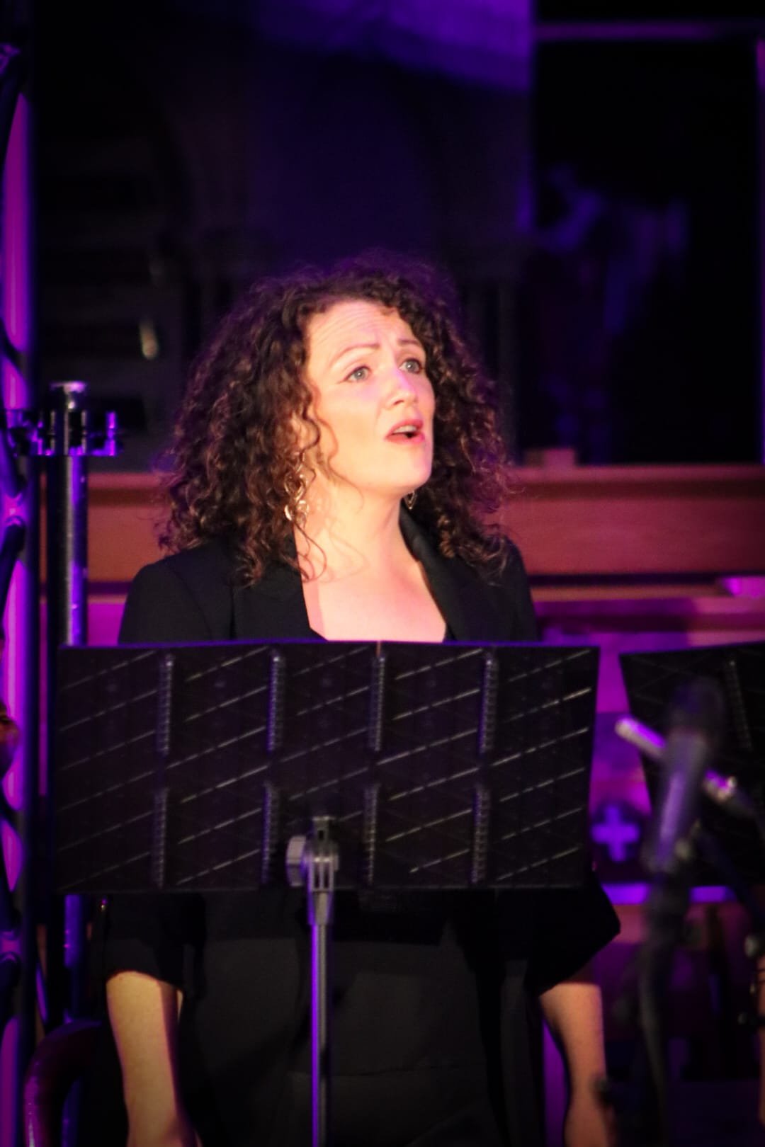 A woman with curly hair singing at a microphone, with music stands and purple stage lighting in the background.
