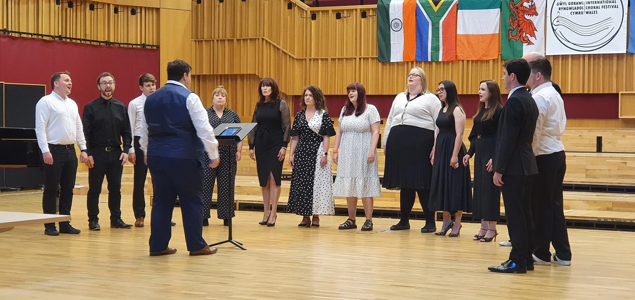A group of people, both men and women, are standing in a semi-circle, singing or performing in a wooden-floored auditorium with flags hanging on the wooden wall behind them.